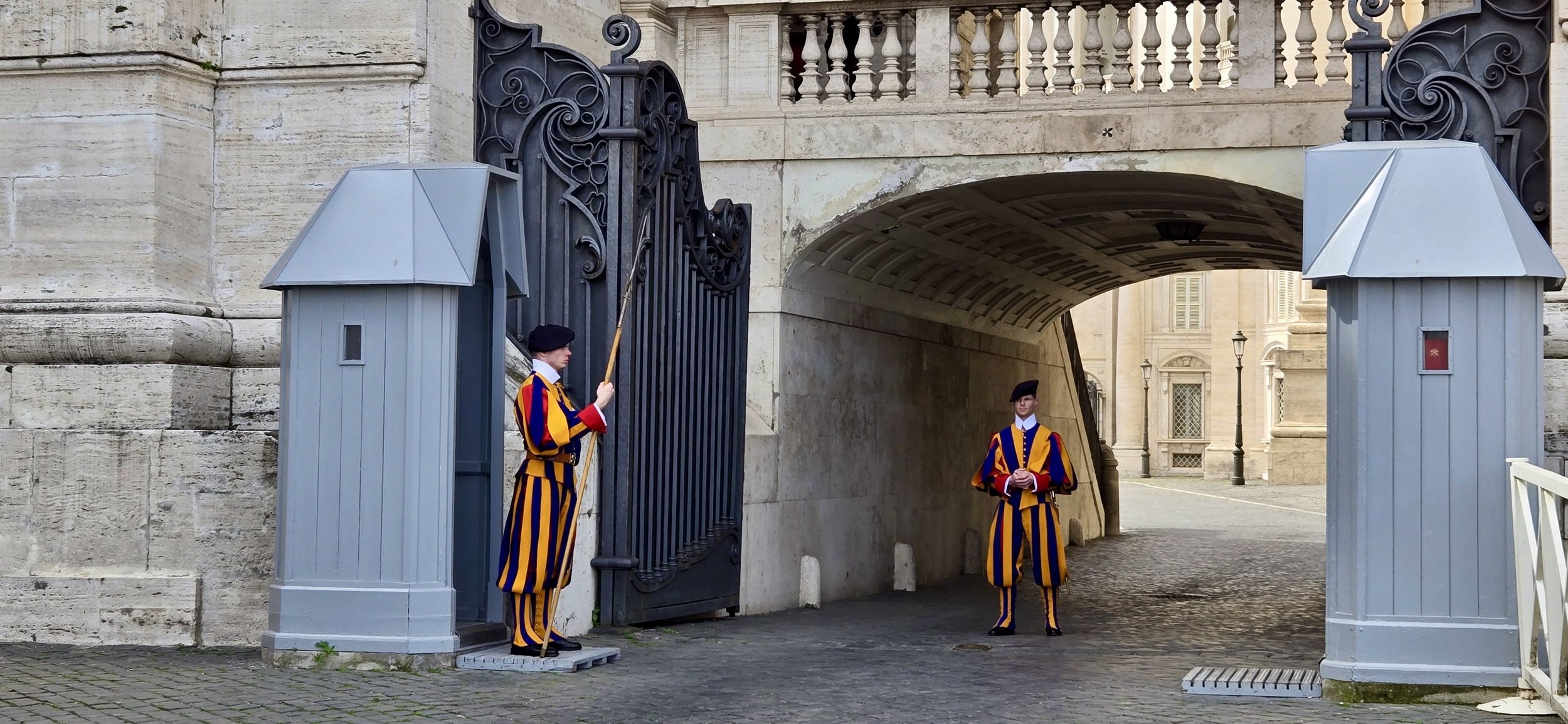 Two guards in colorful striped uniforms and black berets stand under an archway at a historic gate; one guard holds a spear while the other stands at attention.