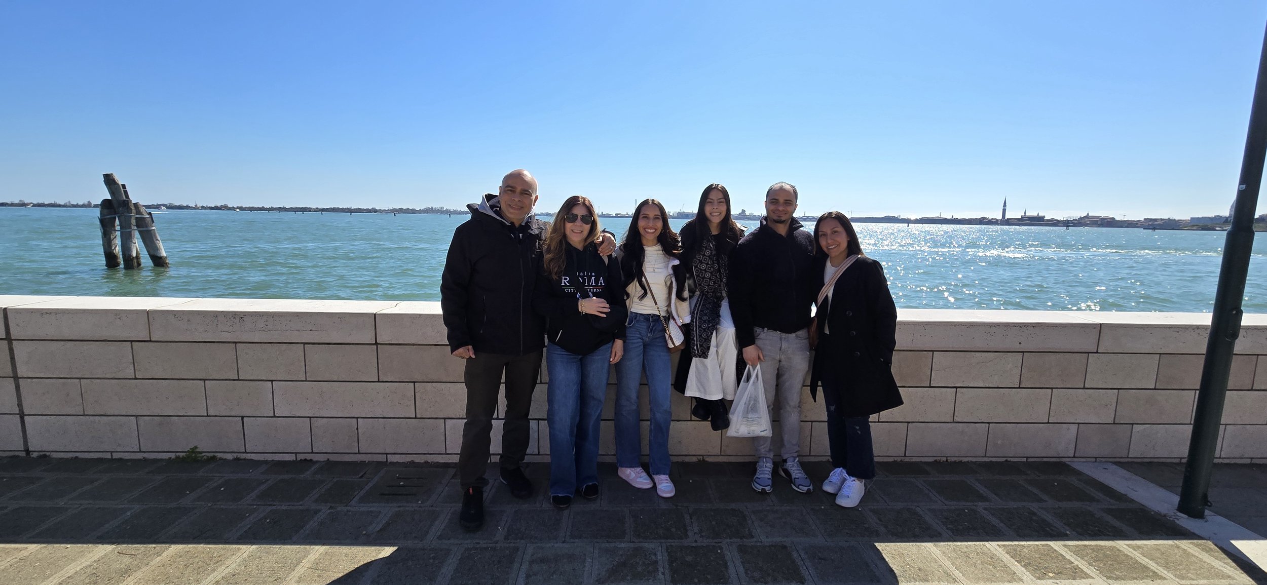 Group of six people standing outdoors by a waterfront on a sunny day, with a body of water and a cityscape in the background.