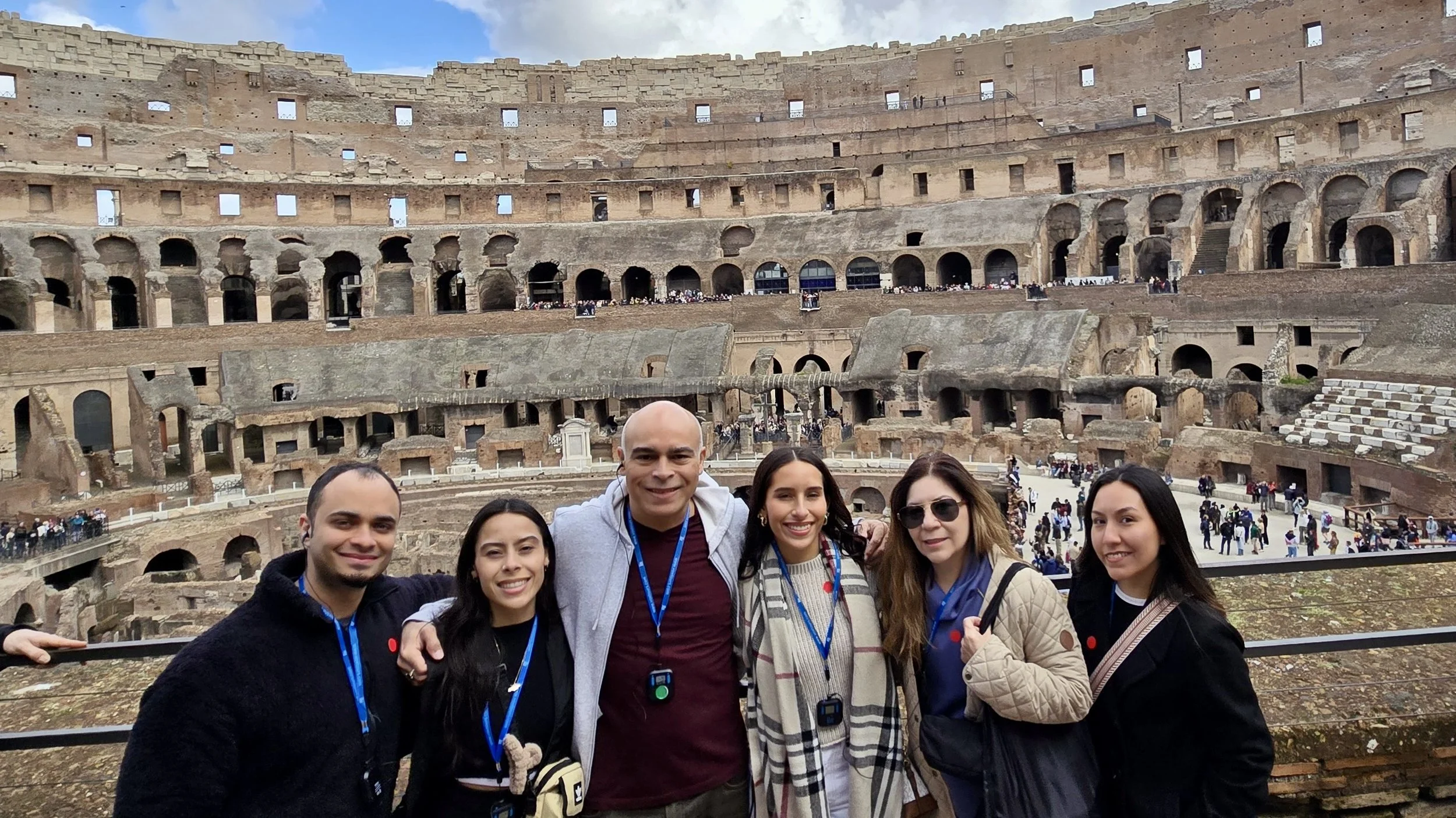 Six people standing together in front of the Colosseum in Rome, Italy, smiling at the camera.