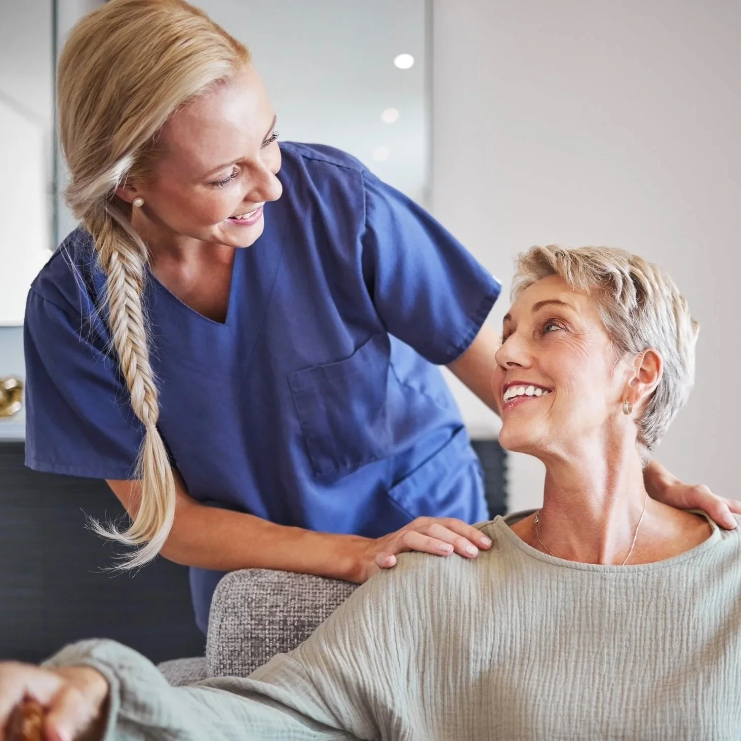 A nurse or caregiver in a blue uniform smiling and talking to a woman, who is sitting and smiling back at her, in a home setting.