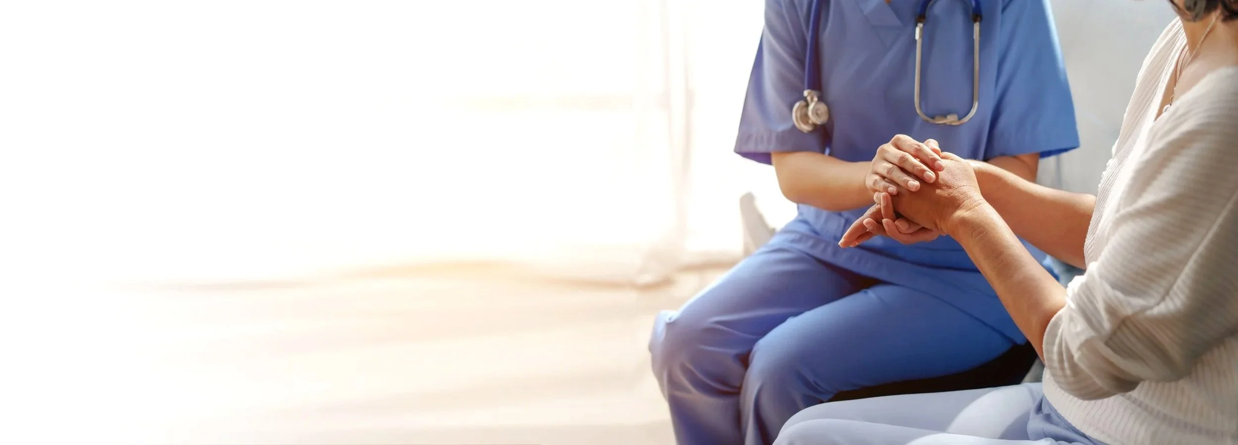 A healthcare professional in blue scrubs holding a patient's hand during a consultation in a bright room.