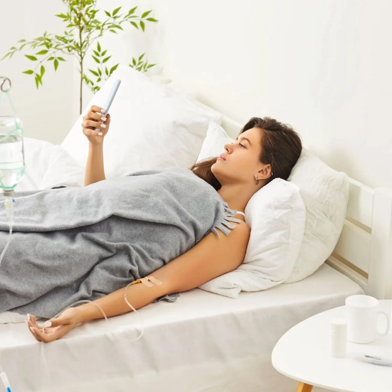 A young woman lying in a bed in her home with an IV, resting and using a smartphone.