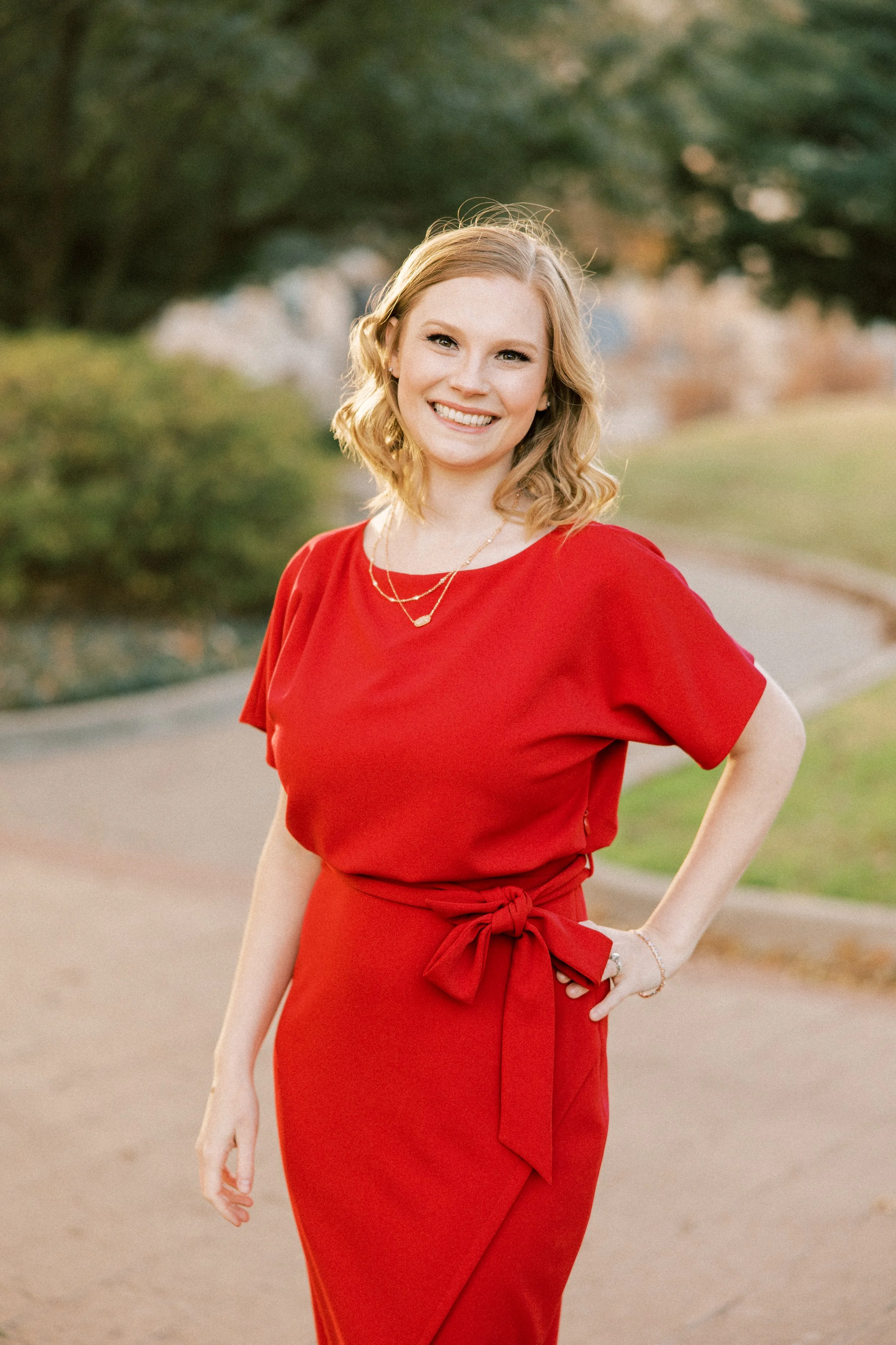 A woman with blonde wavy hair smiling outdoors, wearing a red dress with a waist tie and layered gold necklaces.