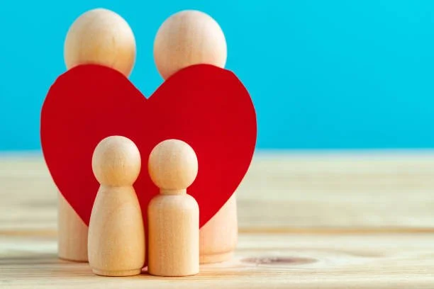 Four wooden peg dolls in front of a red heart shape on a wooden surface with a blue background.