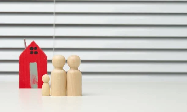 Wooden figurines representing a family of three, with a small red wooden house in the background, against a white surface and horizontal blinds.