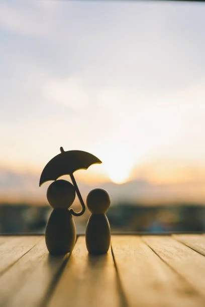 Two small figurines, one holding an umbrella over the other, with a sunset background.