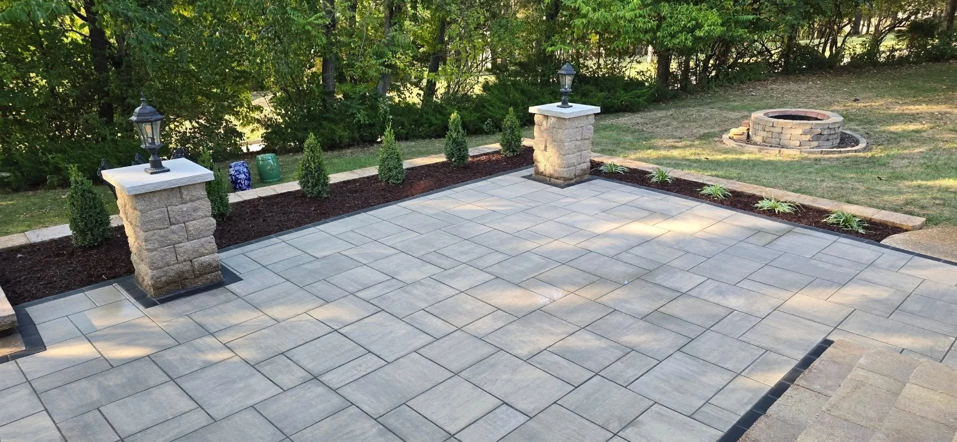 Backyard patio with stone tiles, surrounded by a garden with small shrubs, decorative planters, and a fire pit in the grass.
