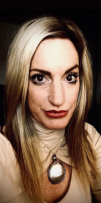 Close-up of a woman with long blonde hair, wearing a beige top and a large pendant necklace.