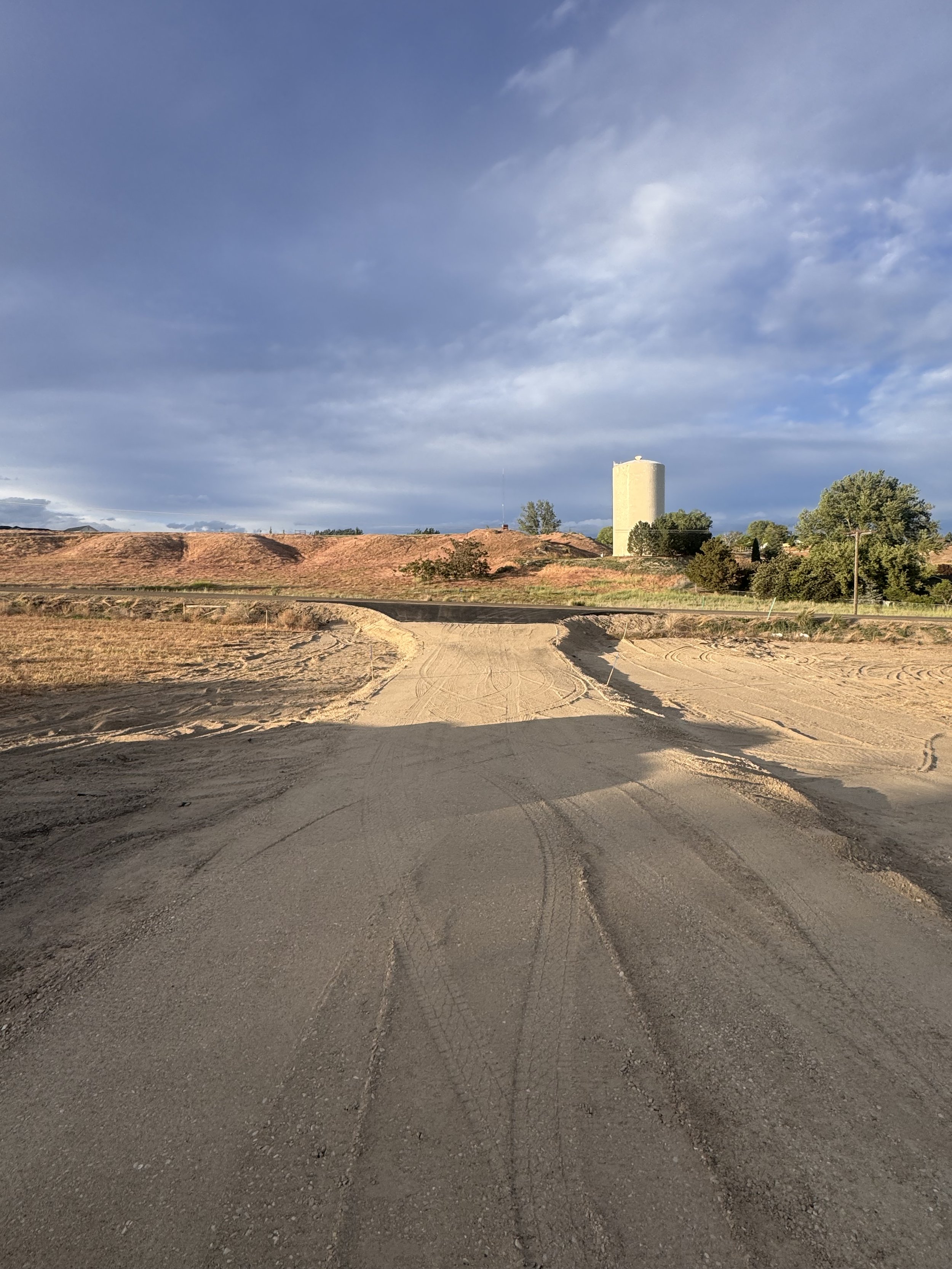 Construction site on a dirt road with a small excavator, workers, and stone barriers, surrounded by trees and a house in the background.