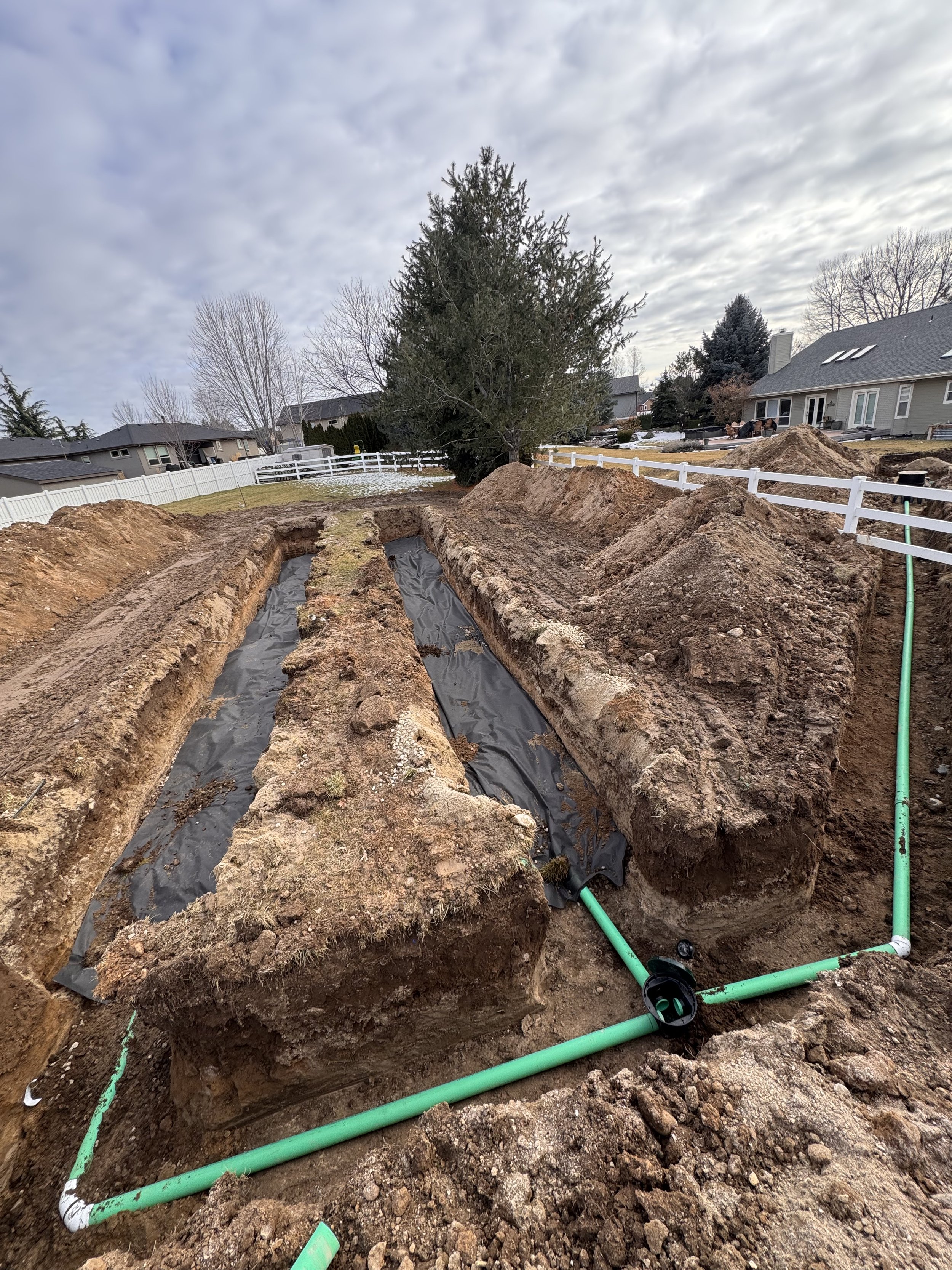 A large hole in the ground showing underground pipes and cables, with a white pipe connected to a black flexible hose, surrounded by soil and some grass near the edges.