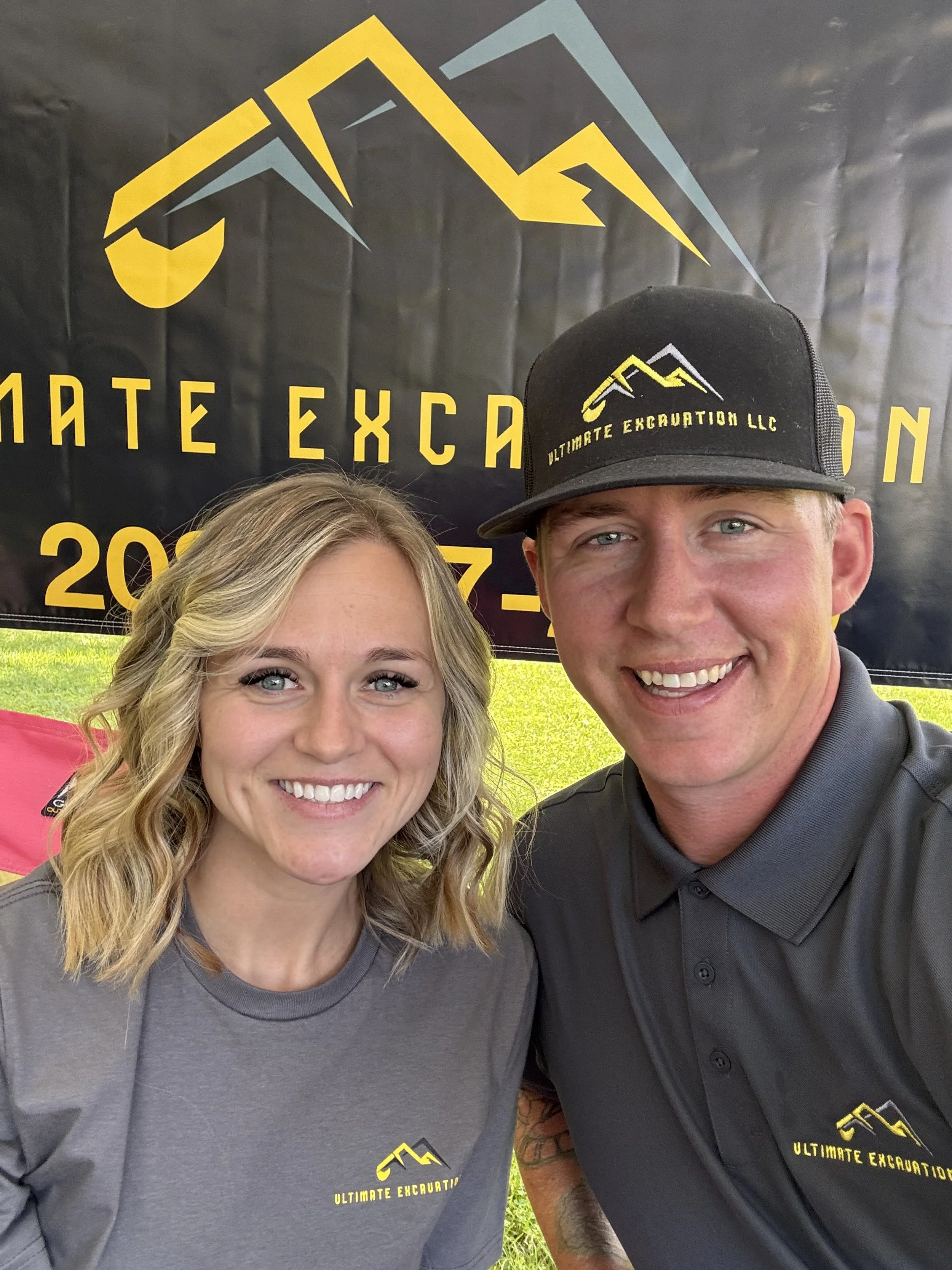 Two smiling people, woman and man, taking a selfie in front of a black banner with yellow and gray mountain logo and text. The woman has shoulder-length blonde hair, and the man is wearing a baseball cap. Both are wearing gray shirts with the same logo and text. The background includes green grass.