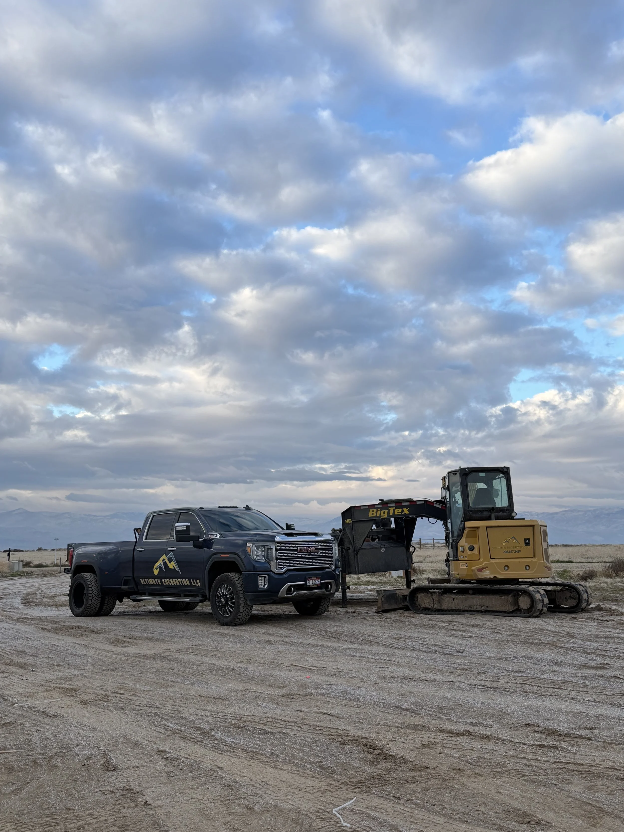 A black GMC pickup truck is parked next to a small yellow excavator on a dirt surface under a cloudy sky, with mountains in the background.