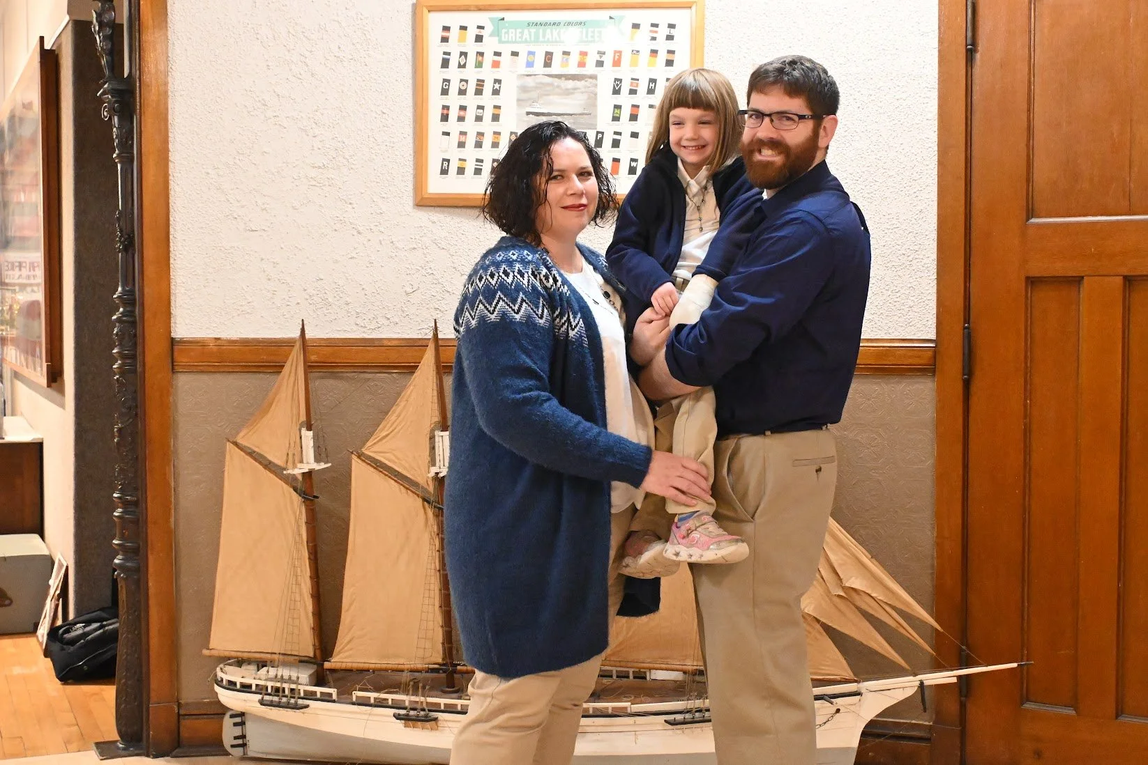 A family of three poses together indoors in front of a decorative model ship. The mother wears a blue patterned cardigan, the father holds a young girl with blonde hair wearing a navy jacket, and the girl is smiling.