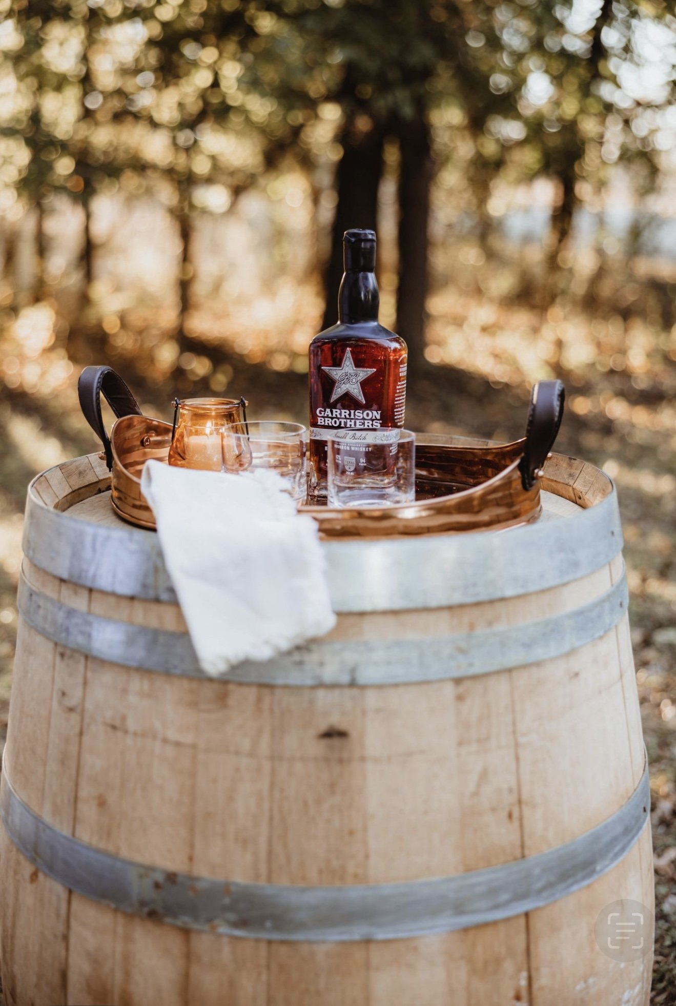 A wooden barrel turned into a table outdoors with a bottle of Garrison Brothers bourbon, two glasses, and a copper tray with a white cloth, set against a background of trees with autumn leaves.