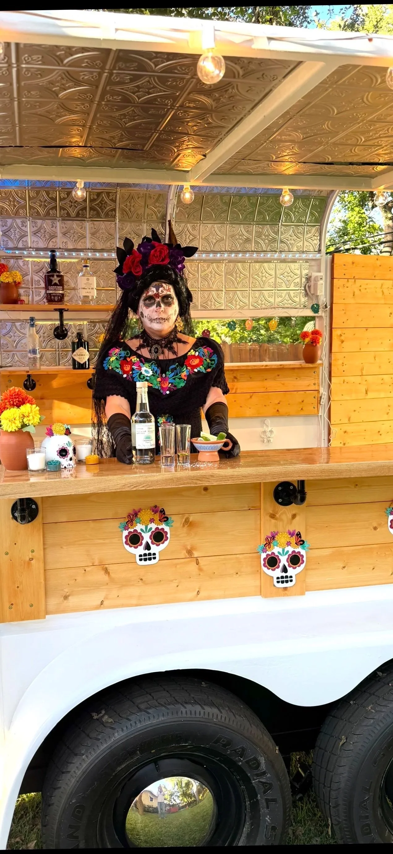 A person dressed in Day of the Dead makeup and traditional Mexican clothing, standing behind a decorated wooden bar truck with floral and skull decorations. The setting appears to be an outdoor daytime event.