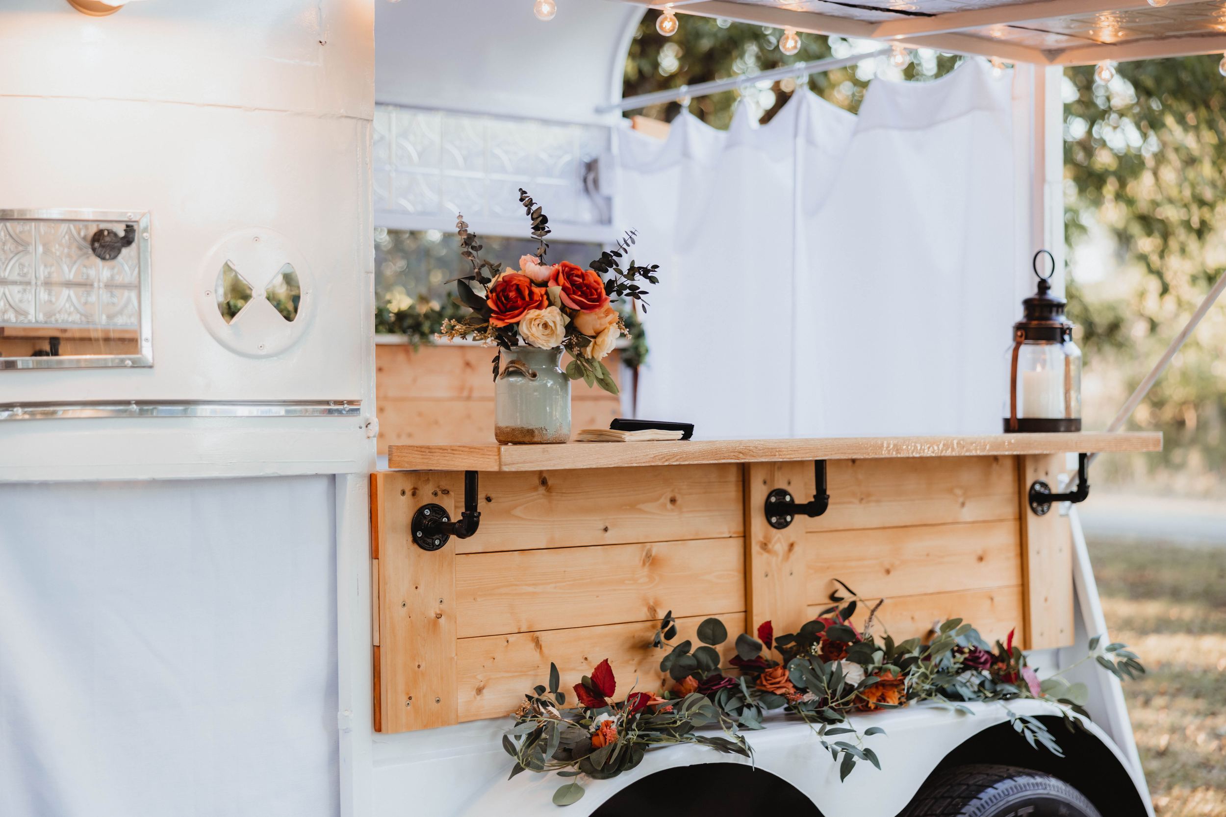 A white food or beverage trailer with a wooden countertop decorated with a bouquet of orange, cream, and pink flowers in a green vase, and a candle lantern. There are string lights above and greenery around.