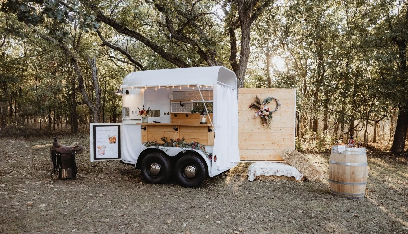 A white food truck with string lights park in a wooded area during sunset. It is decorated with a wooden panel and floral wreath, with hay bales and a barrel nearby.