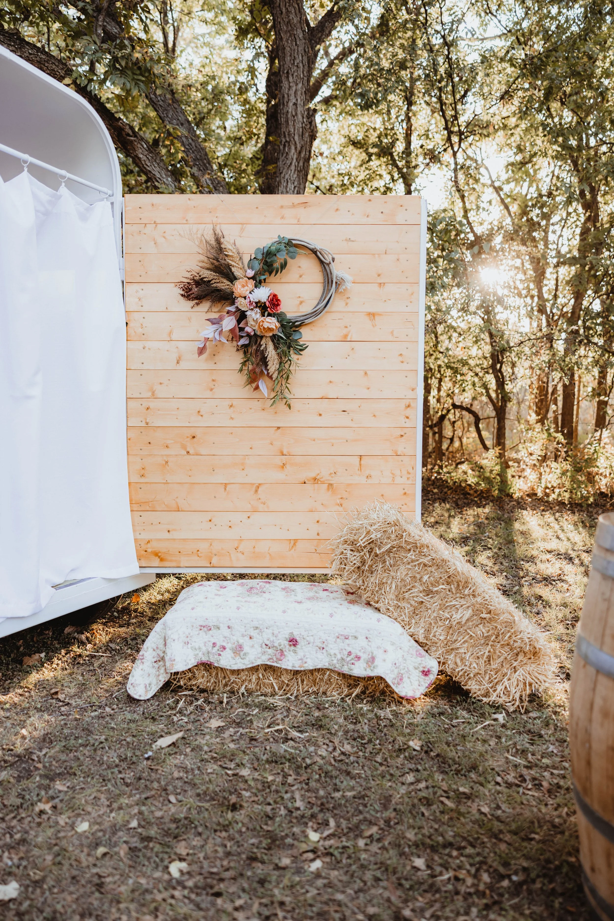 A rustic outdoor decor scene featuring a wooden backdrop decorated with a floral wreath, a hay bale with a floral cloth, and a hay cushion in a forest setting during sunset.