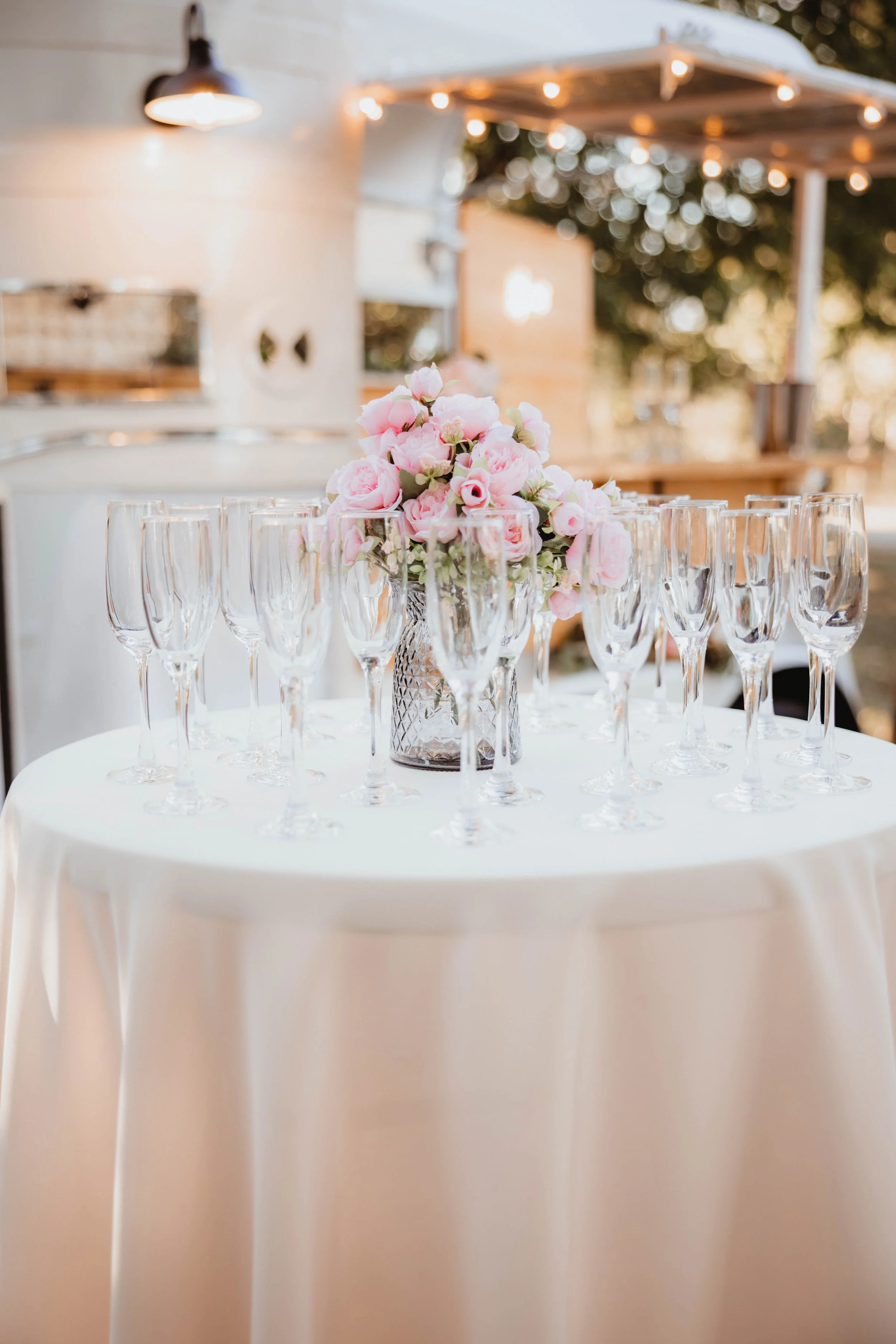 A table with a white tablecloth set with multiple empty champagne flutes and a glass vase with pink and white flowers, outdoors in a decorated setting with string lights and a wood structure in the background.