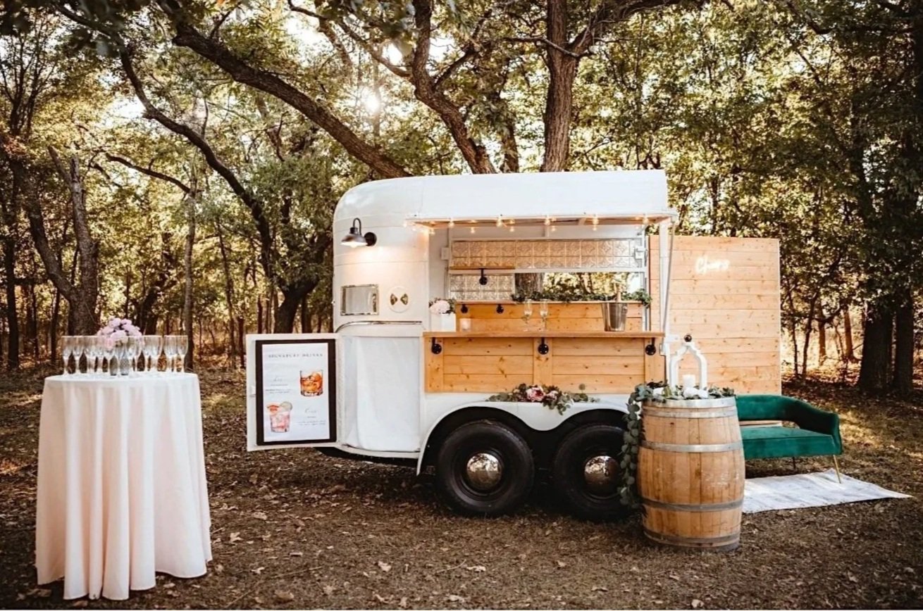 A vintage white food truck with a wooden serving window, set outdoors in a wooded area with tall trees and sunlight filtering through. To the left, a tall round table draped in a white tablecloth holds glassware and a flower arrangement. To the right, a wooden barrel with floral decoration serves as a stand, next to a green velvet sofa with a striped rug underneath.
