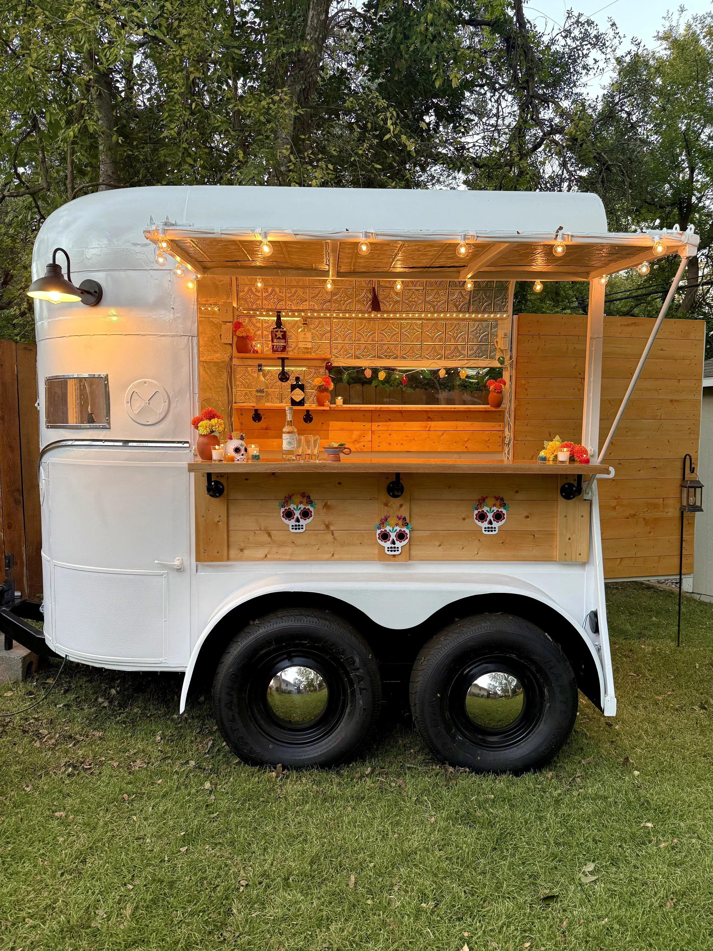 A mobile bar setup inside a white vintage trailer with wooden accents, decorated with hanging string lights and colorful skull decorations, set outdoors on a grassy area with trees in the background.
