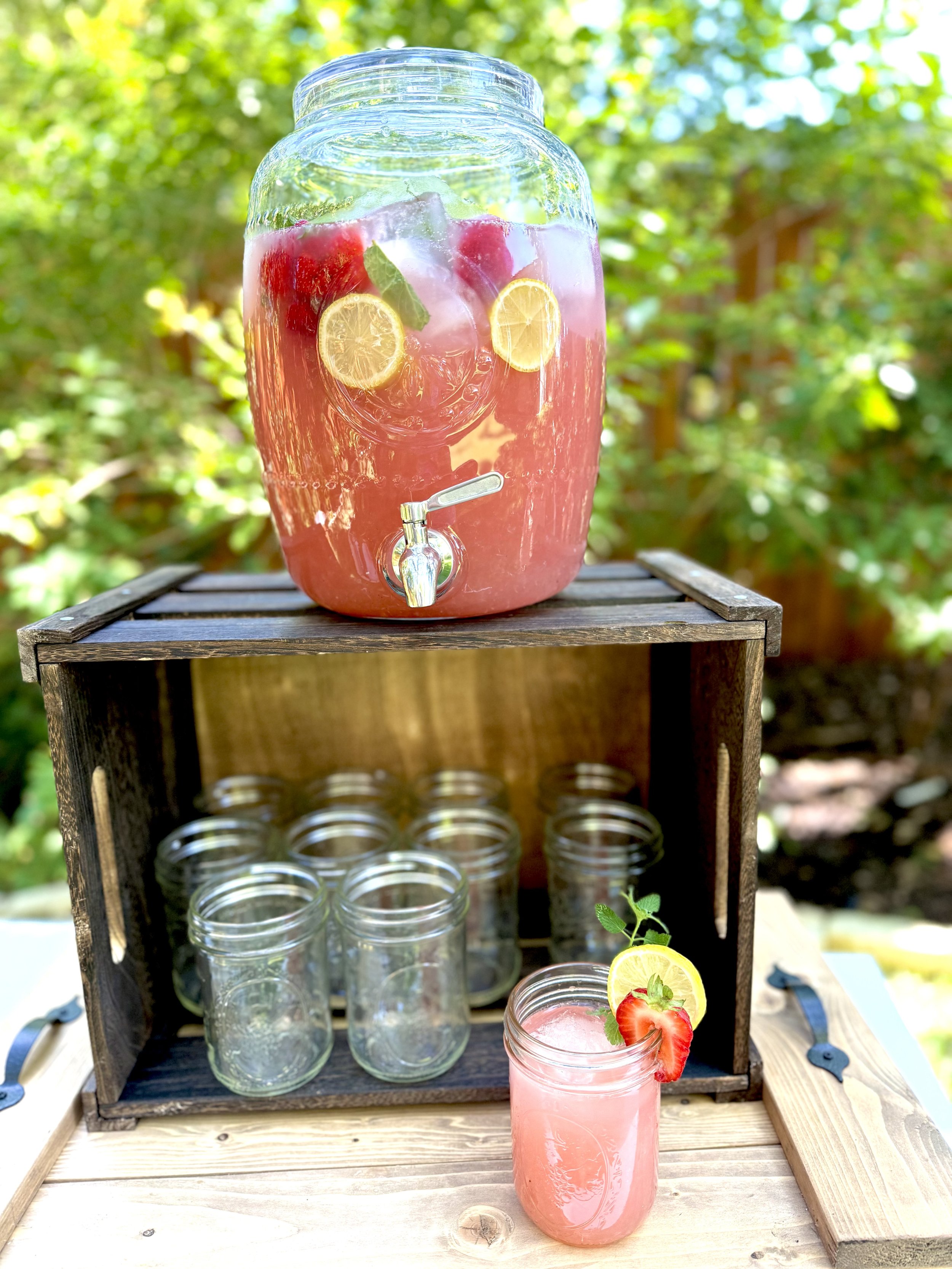 A glass beverage dispenser filled with pink strawberry lemonade, slices of lemon, strawberries, and mint leaves, sitting outdoors on a wooden table. Below the dispenser is a wooden crate holding empty jars, with a mason jar of lemonade garnished with a lemon slice, strawberry, and mint leaves placed in front.