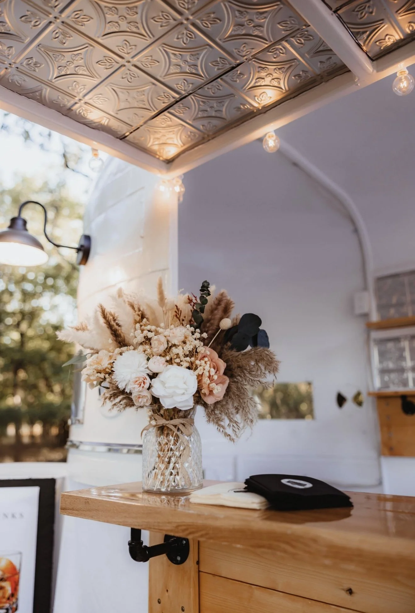 A floral arrangement with white, pink, and peach flowers in a textured glass vase on a wooden counter inside a decorated space with string lights and a white wall.