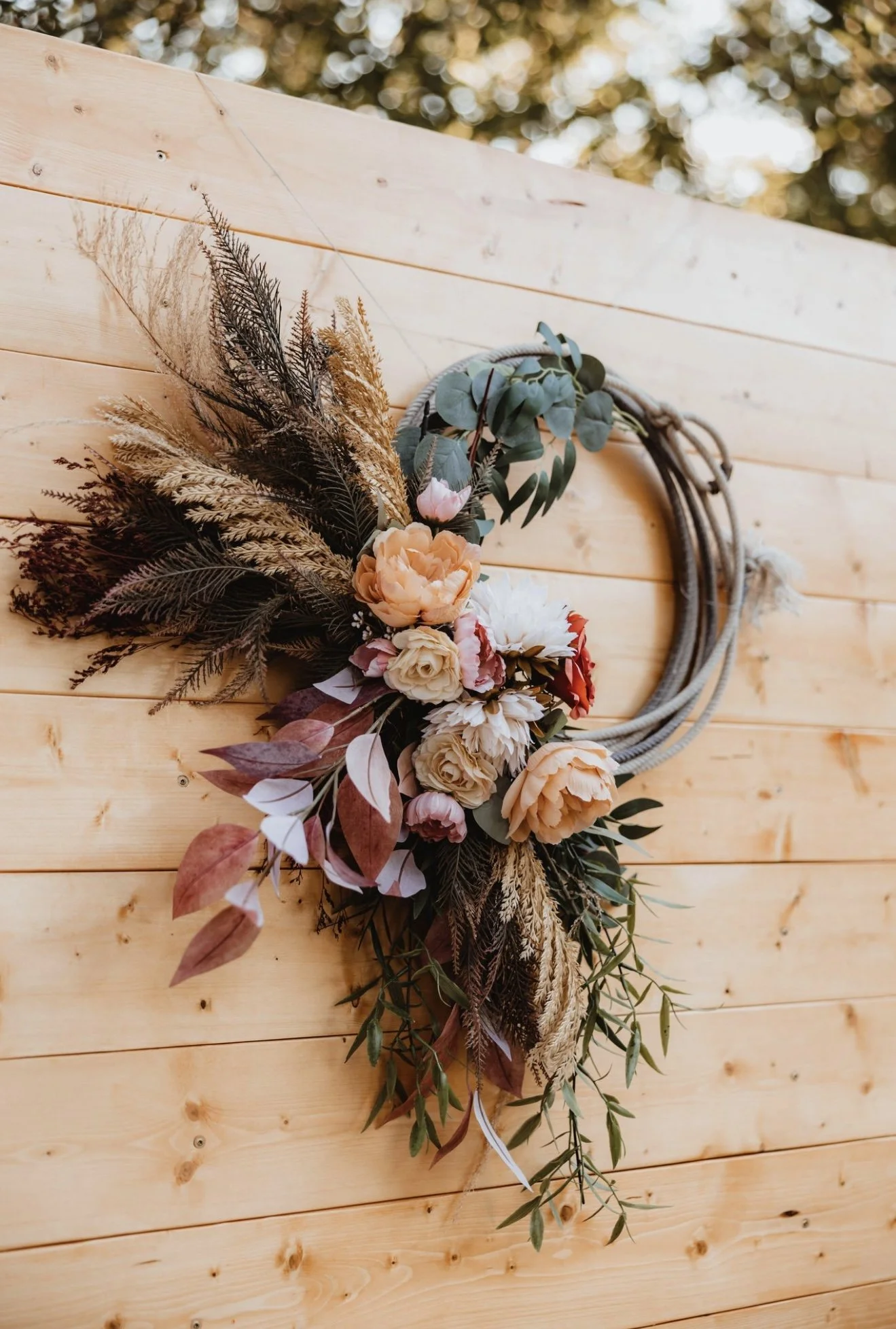 A floral wreath with peach, white, and pink flowers, dried grasses, and green foliage hanging on a light wooden wall.