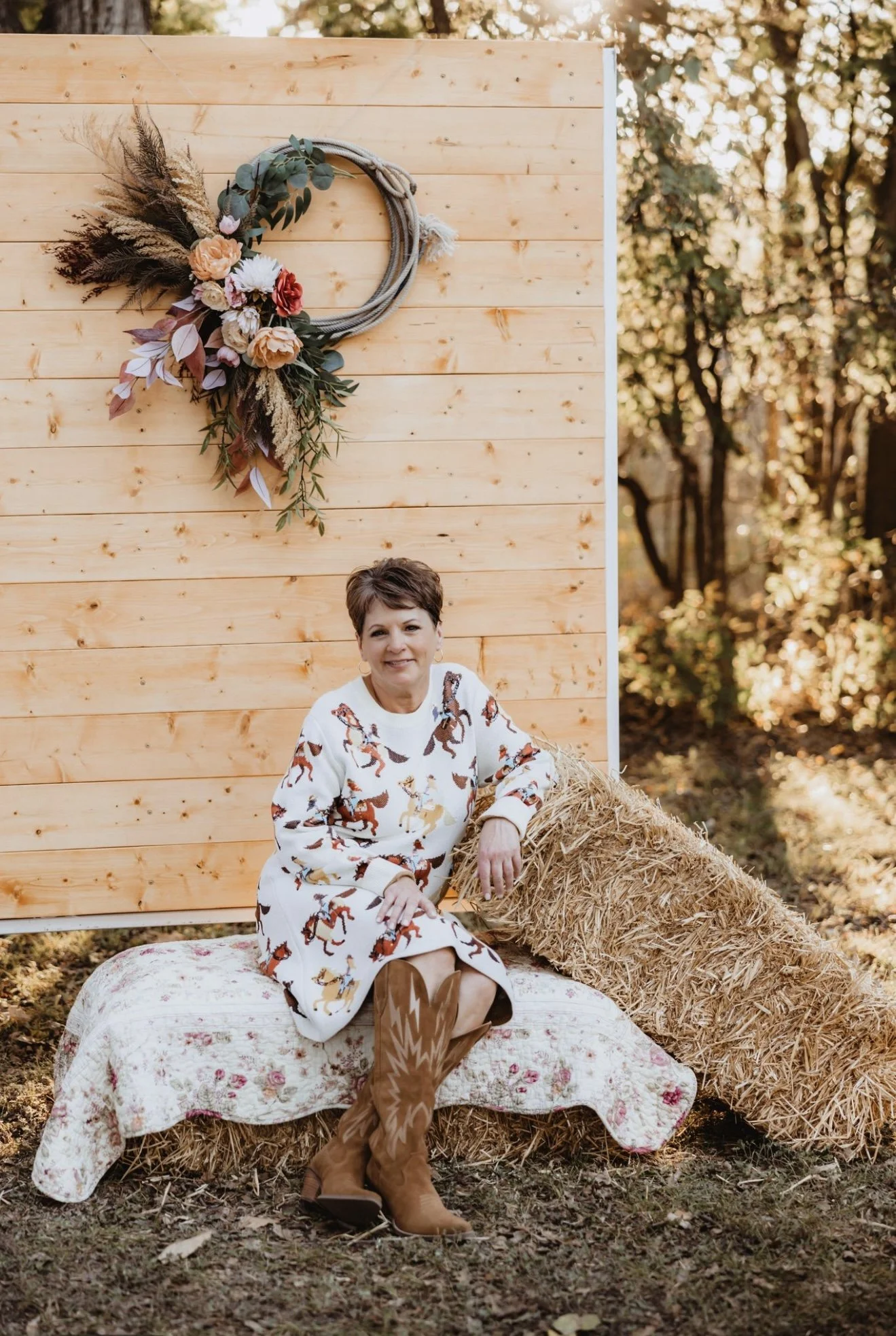 A woman sitting on a hay bale outdoors in front of a wooden backdrop decorated with a floral hoop wreath. She is wearing a white dress with a horse pattern, brown cowboy boots, and has short dark hair. There is a floral quilt draped over the hay bale