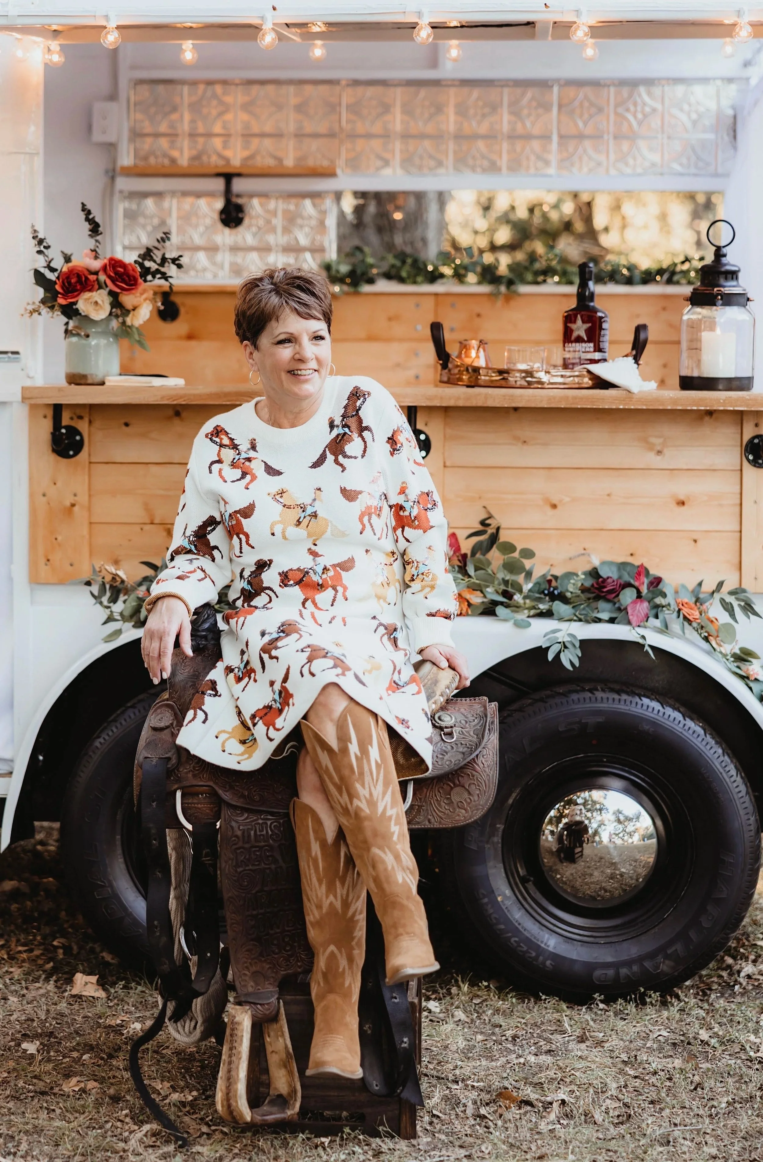 A woman sitting on a saddle in front of a decorated food truck at an outdoor event, wearing a white dress with horse prints and tan cowboy boots.
