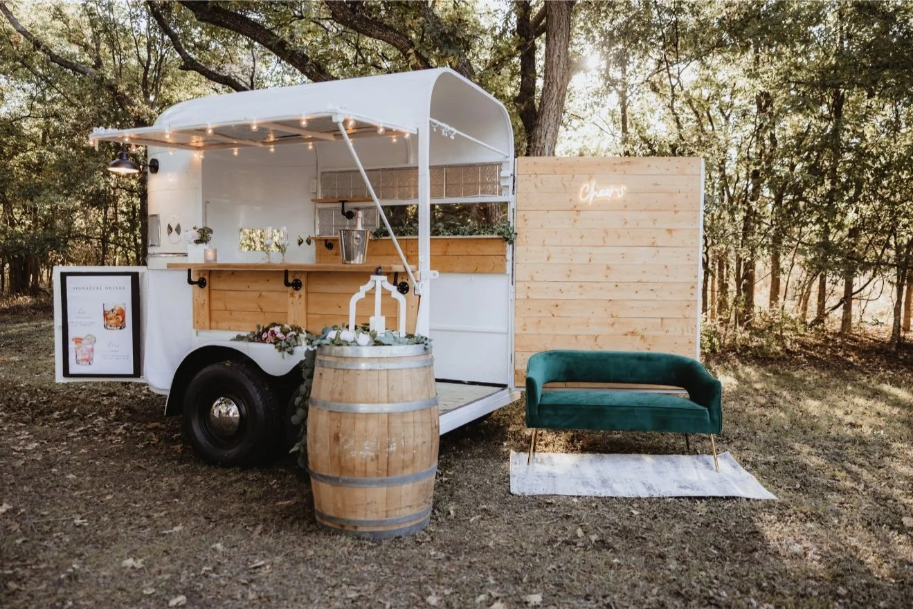 A food truck with a white and wooden exterior is set up in a wooded outdoor area, decorated with string lights, greenery, and flowers, with a barrel in front, a teal velvet loveseat on a small white rug nearby, and a small sign displaying drink optio