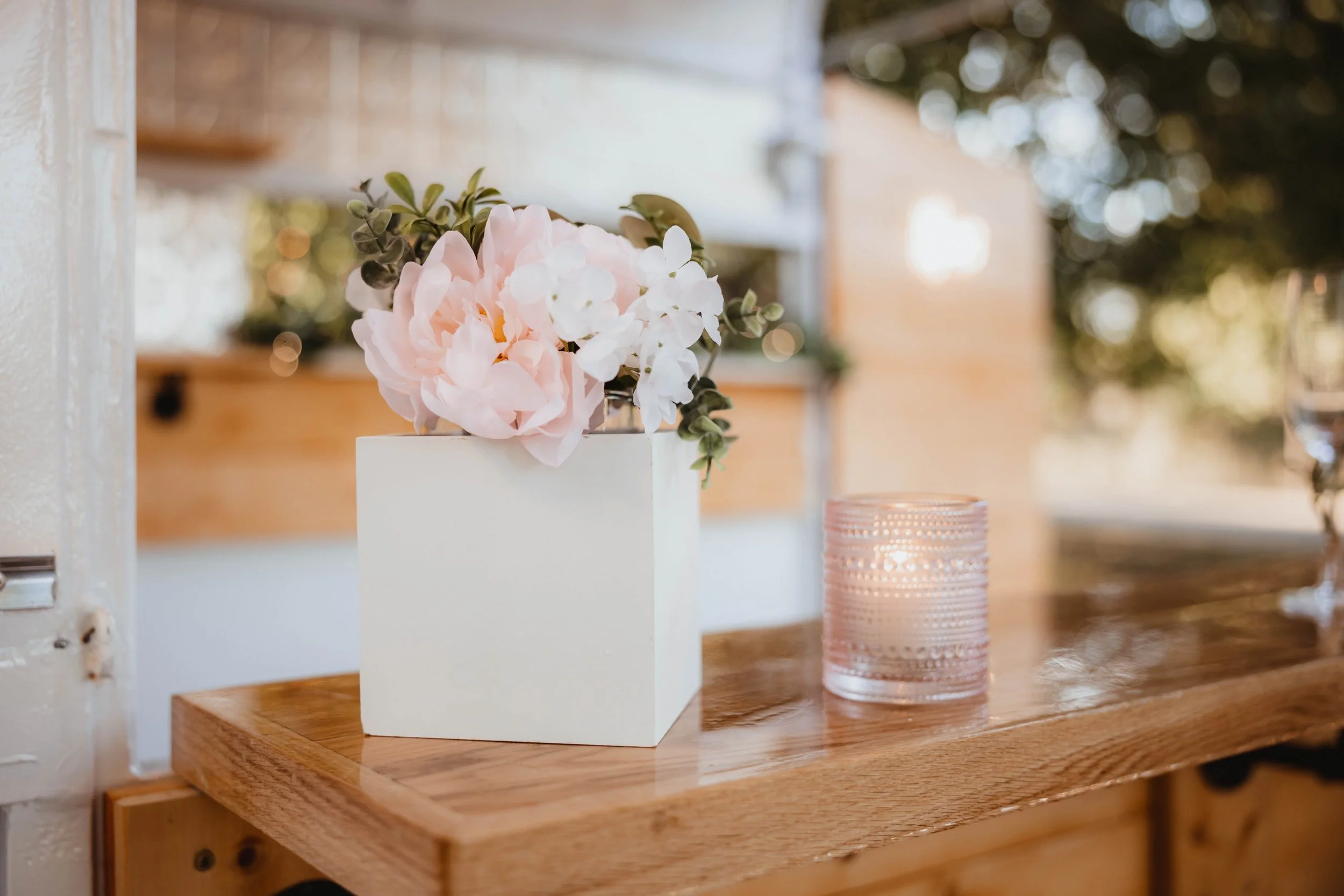 Close-up of a white square vase with pink and white flowers on a wooden surface with a glass candle holder nearby.