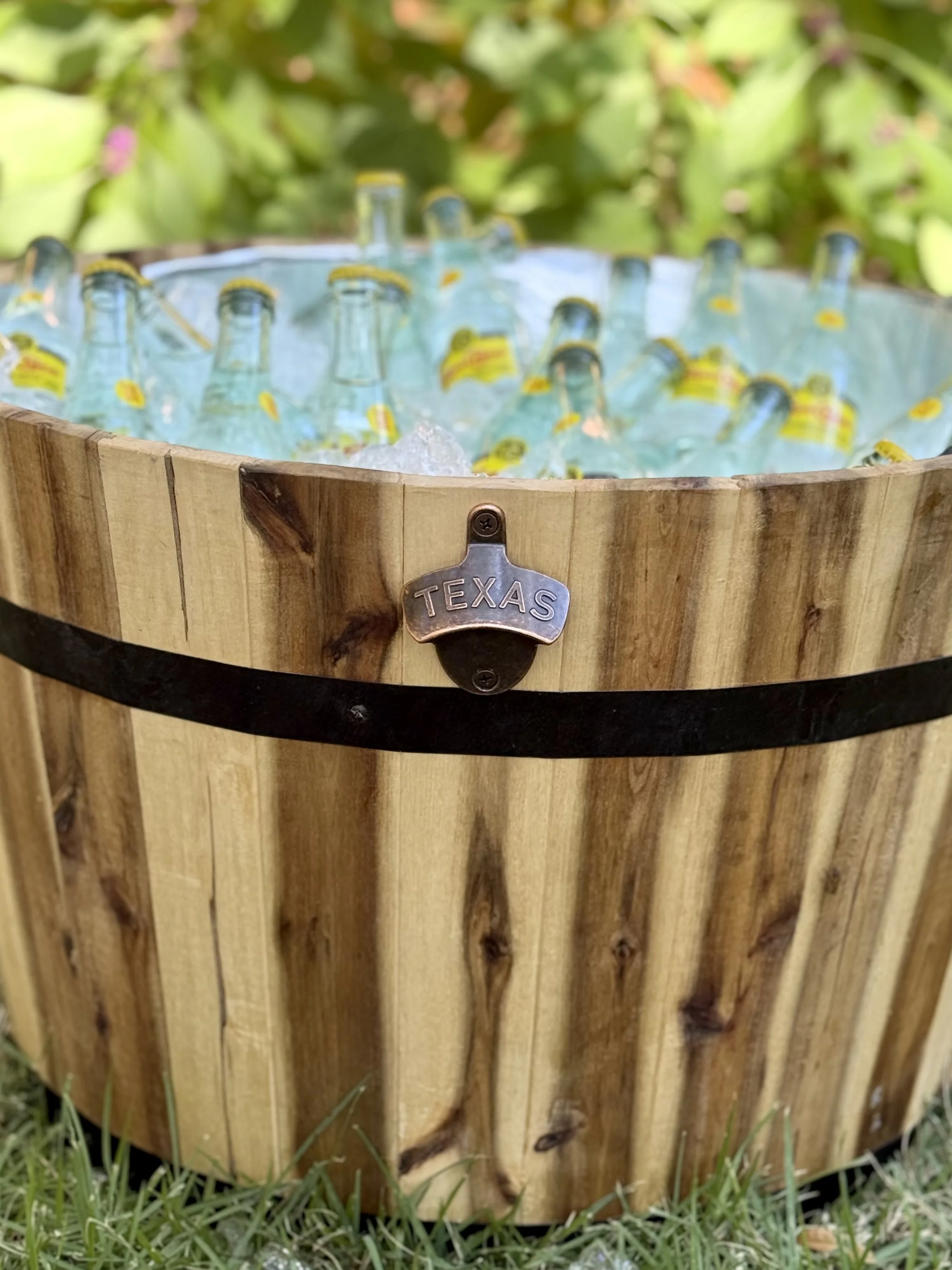 A rustic wooden ice bucket filled with ice and empty glass bottles branded with a yellow and red label. The bucket has a metal badge labeled 'TEXAS' attached to its front, and it is situated on grass with leafy background.
