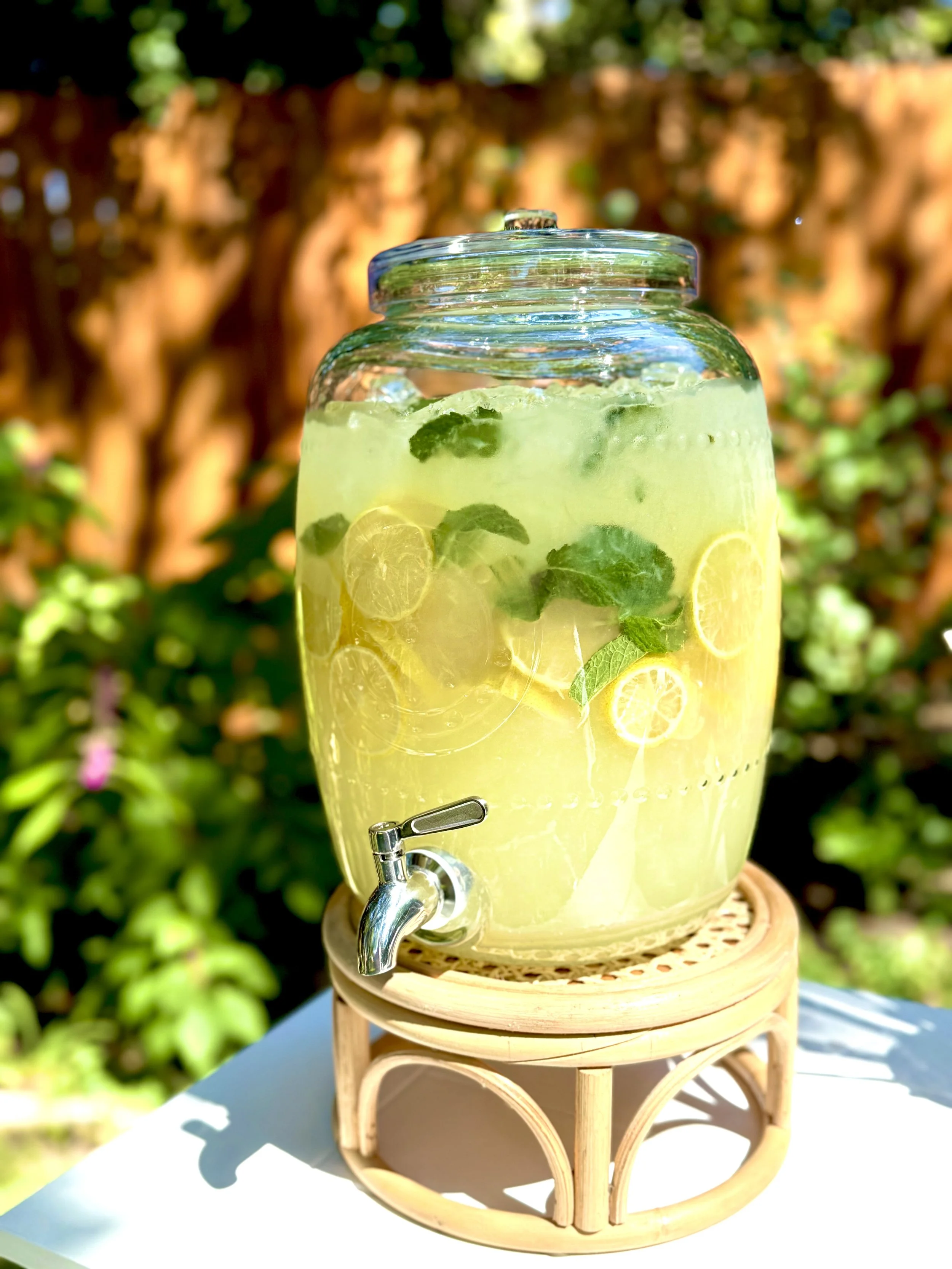 Large glass jar of lemonade with lemon slices and mint leaves, on a wooden stand outdoors with a blurred garden background.