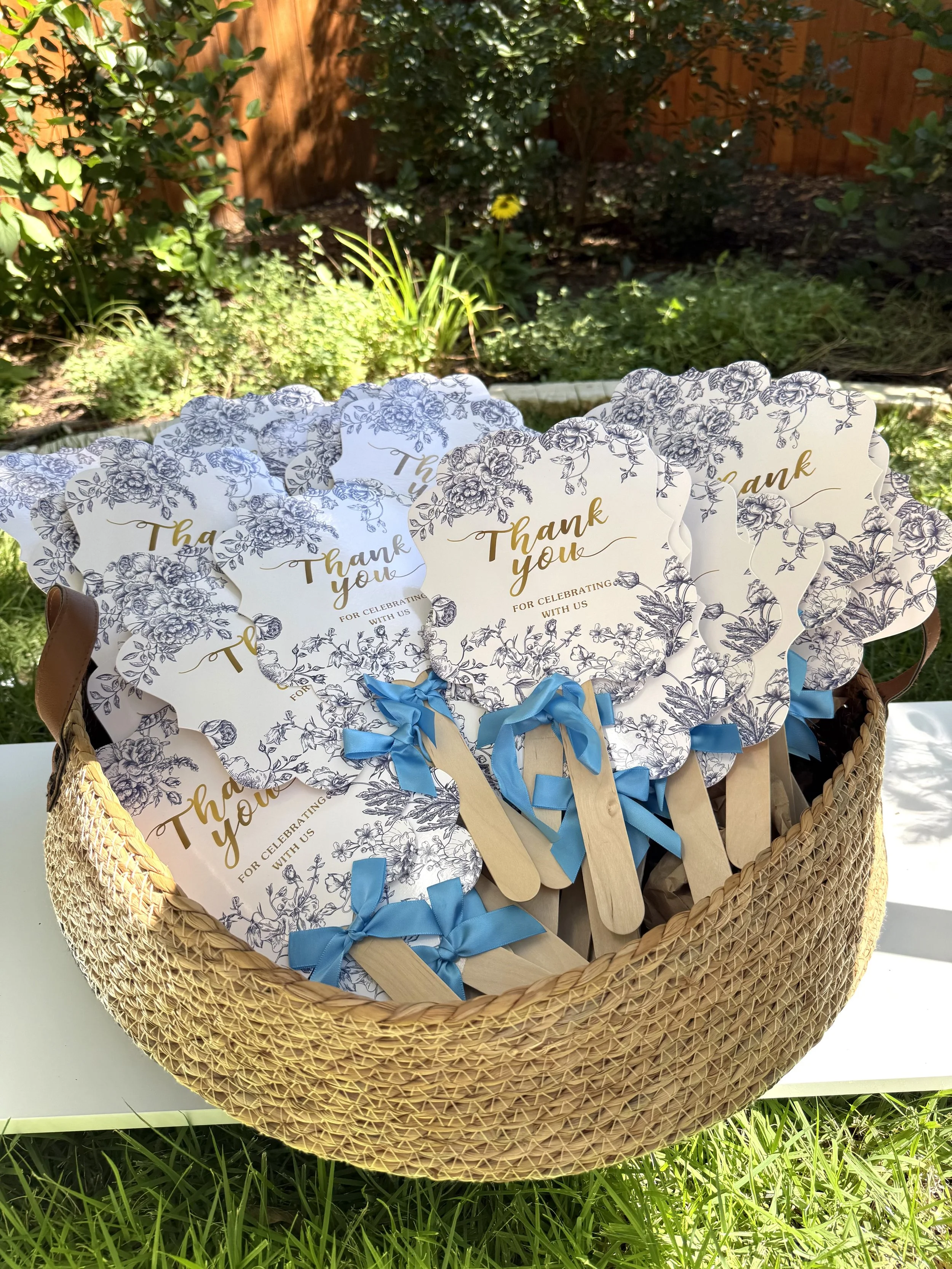 A wicker basket filled with thank you handheld fans with blue ribbons, set outdoors on a white bench, with greenery and a wooden fence in the background.