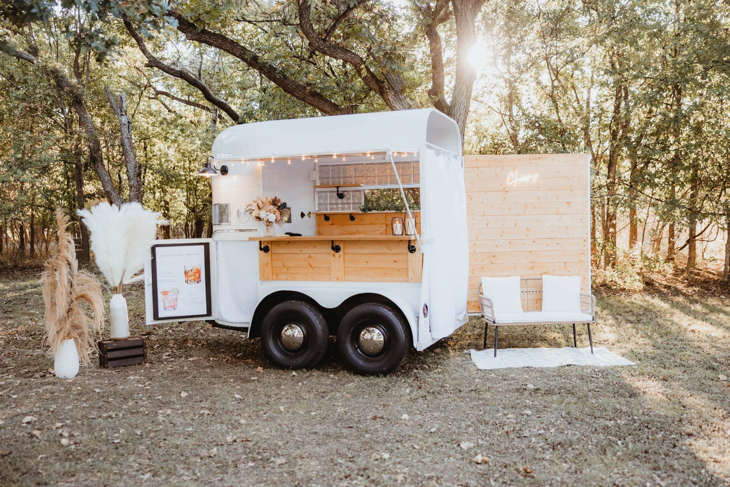 A white food truck with a wooden counter and a small porch is in a forested area with sunlight filtering through the trees. To the left, there is a white vase with pampas grass and a small black crate, and to the right, a white bench with pillows pla