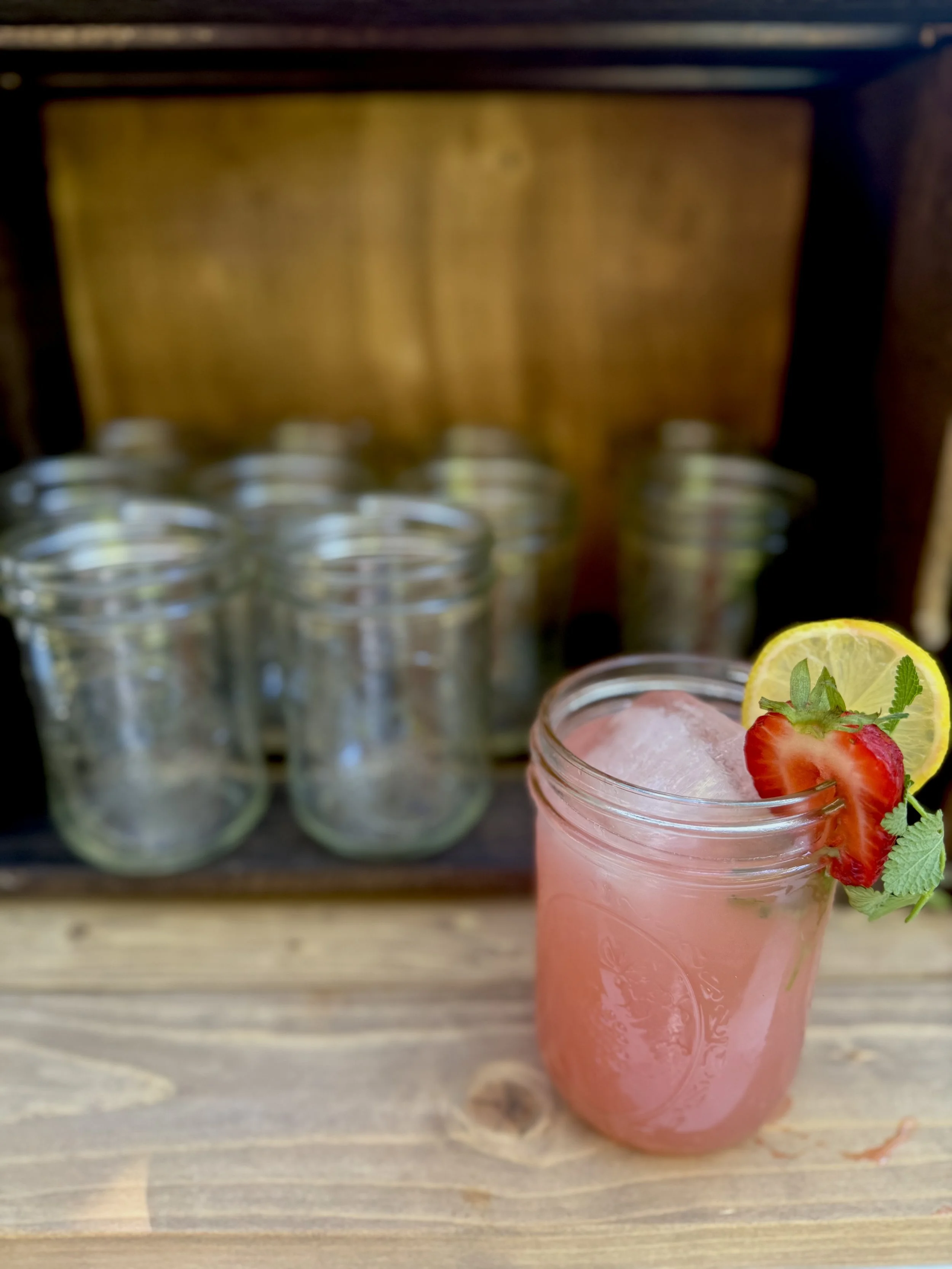 A pink beverage in a mason jar with lemon, strawberry, and mint garnish on a wooden surface, with empty jars and a wooden box in the background.