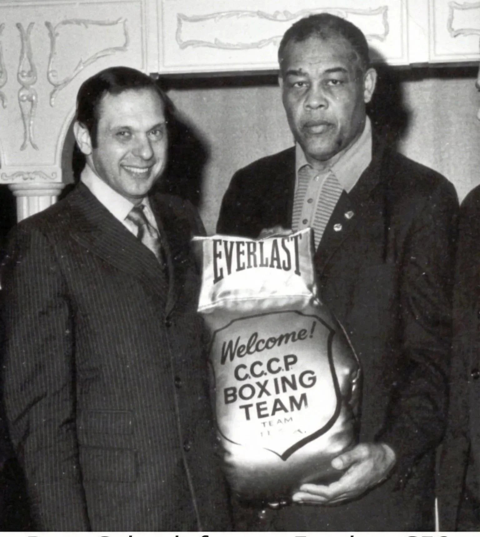 Two men, one with a striped suit and the other with a dark suit, stand together holding a large sack with the words "Welcome! C.C.P. Boxing Team" and a boxer's brand logo. The left man is smiling, and the right man has a neutral expression. The background features decorative wall molding.