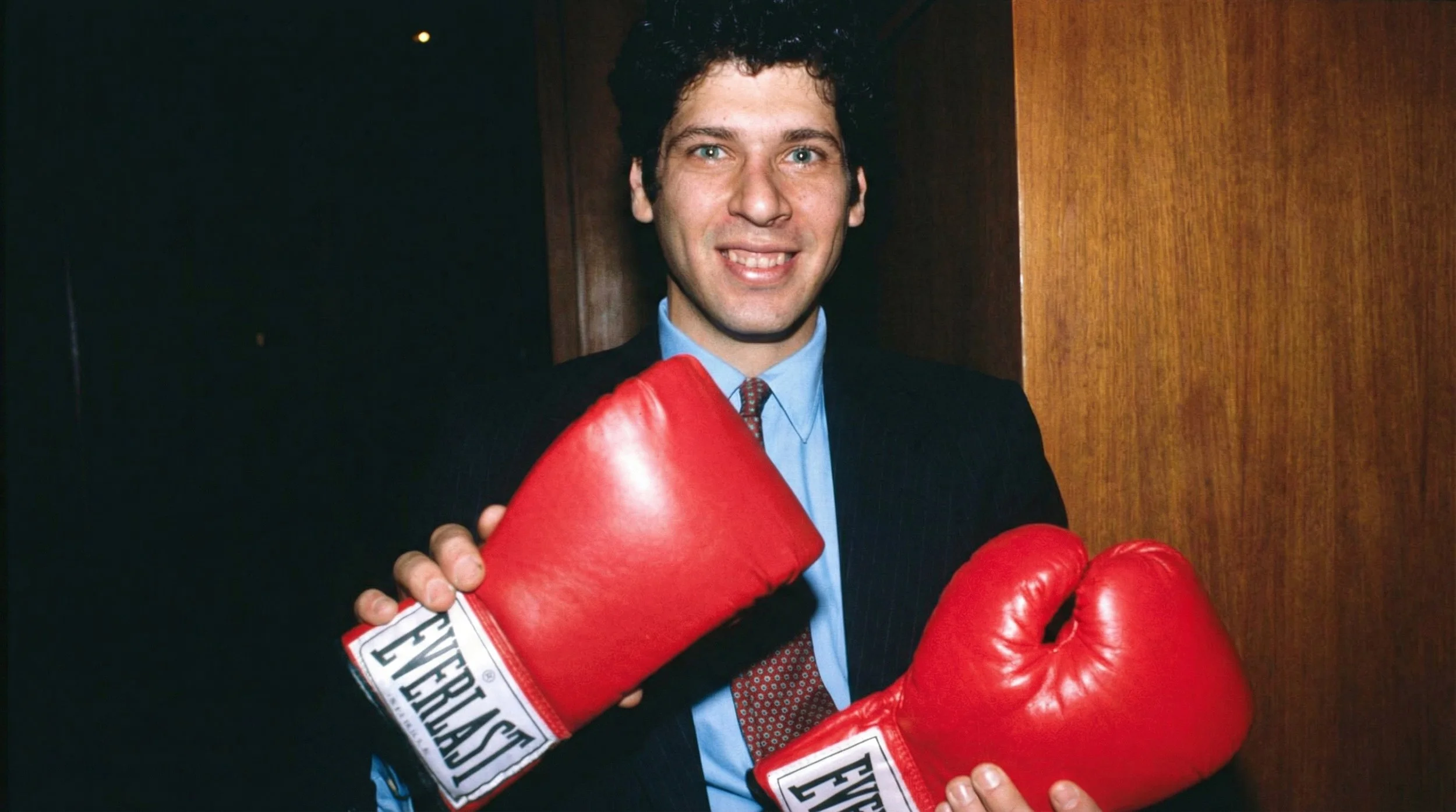 A man in a suit and tie smiling and wearing red boxing gloves.
