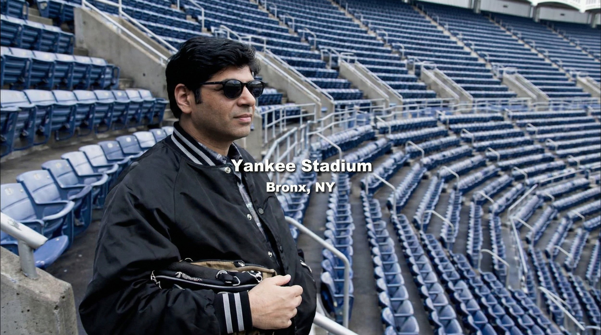 A man wearing sunglasses and a black jacket standing in empty blue seats at Yankee Stadium in Bronx, NY.