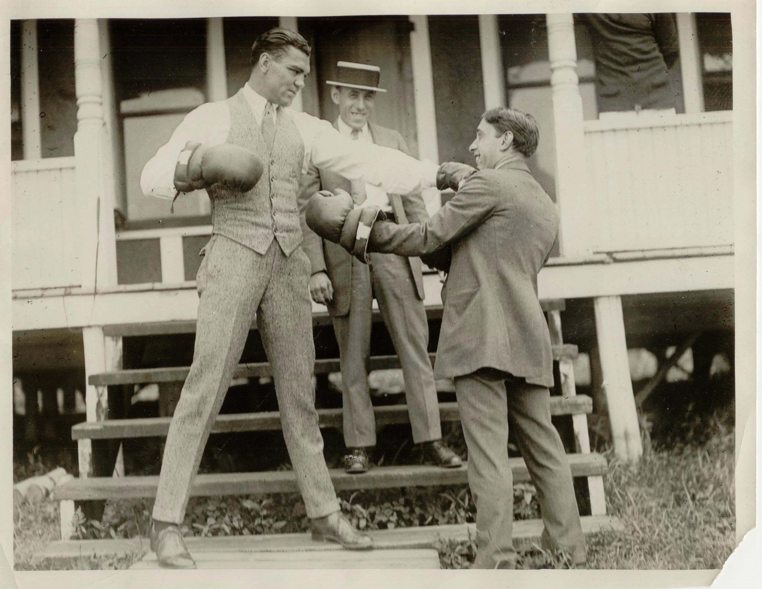 Vintage black and white photo of three men in suits, with two wearing gloves and boxing gear, on a porch with stairs. One man appears to be sparring while the others watch.