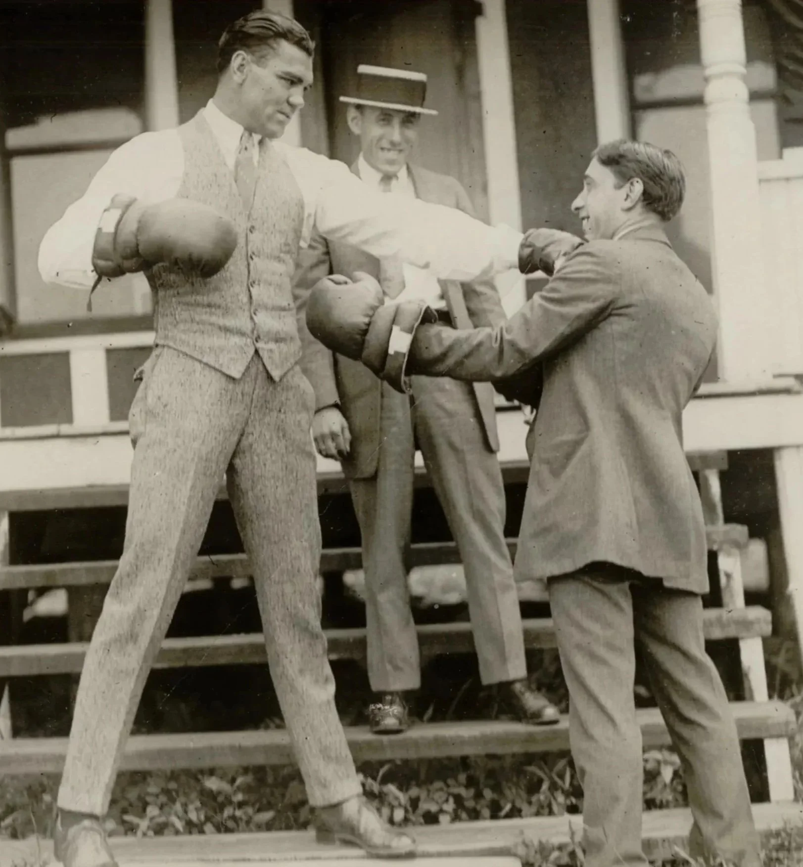 Vintage black and white photo of three men in suits with boxing gloves; one man on the left is throwing a punch at another man on the right, who is partially bent, while the third man in the background watches.