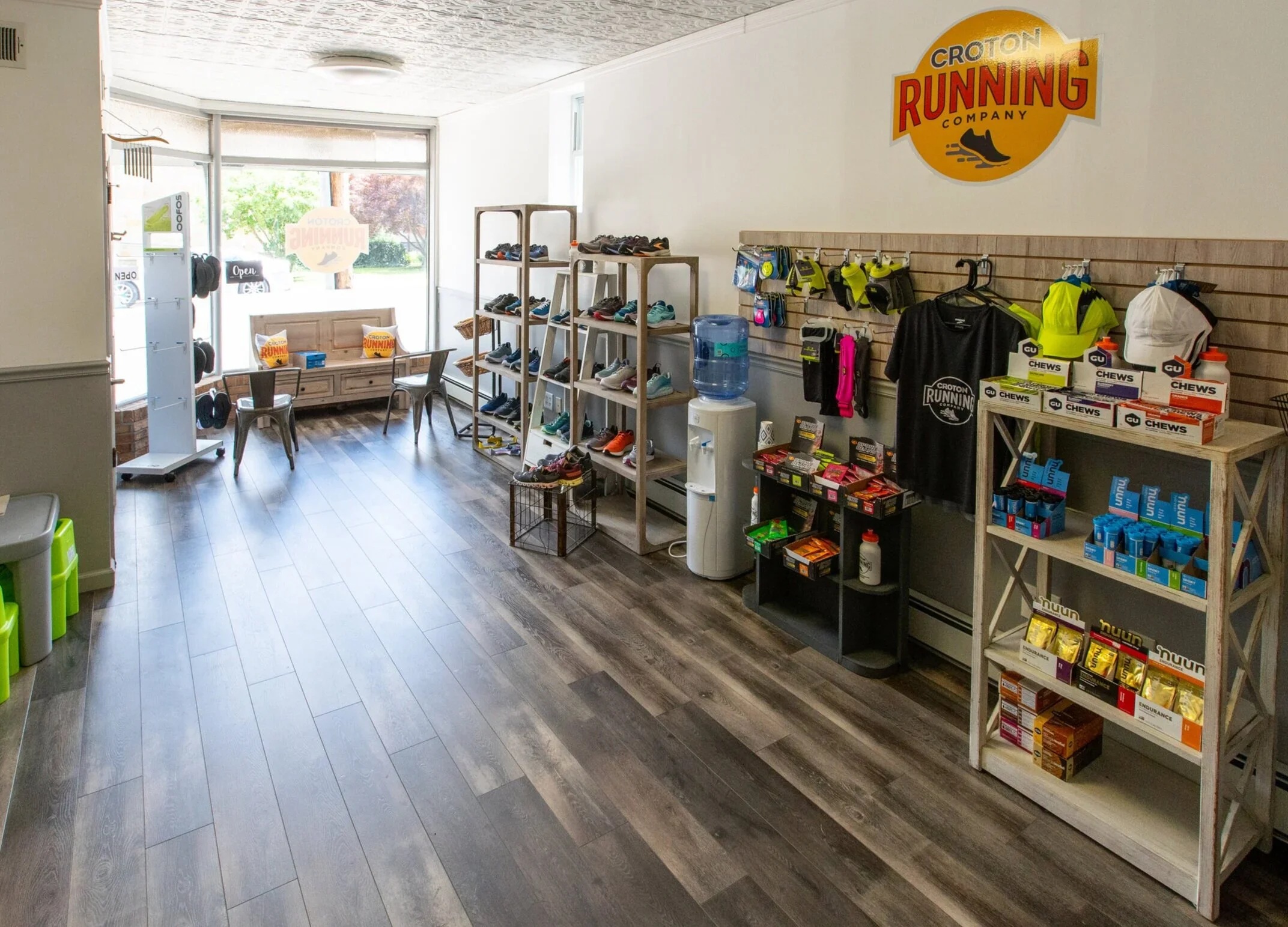 Inside a running shop with shoes on display, a water dispenser, and running gear, with a large window at the front letting in natural light.