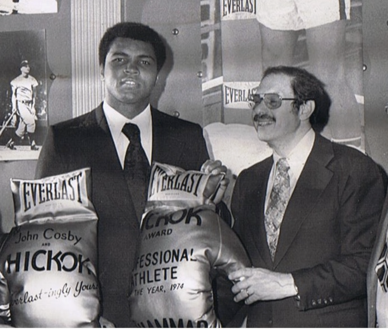 Black and white photo of a young man in a suit and tie, receiving an award from a man with glasses and a mustache, both standing in front of boxing memorabilia and bags marked 'Everlast'.
