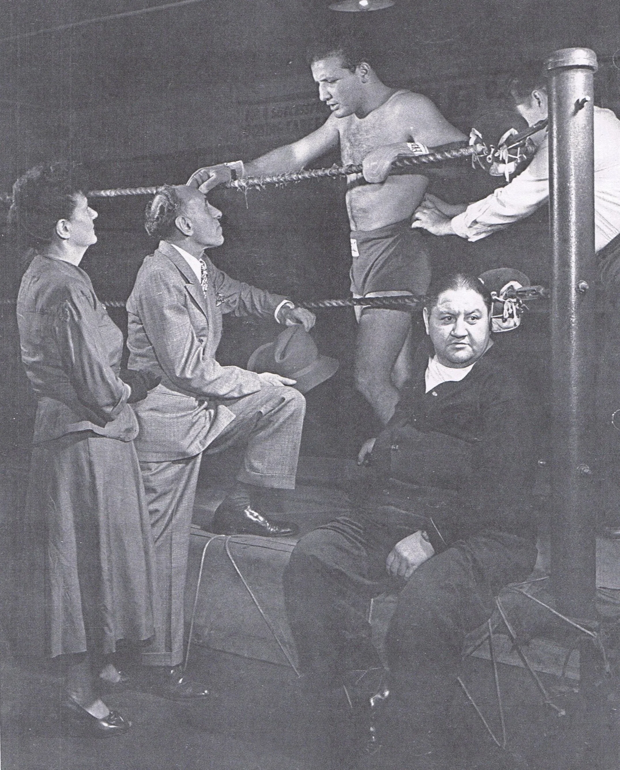 Black and white photo of a boxing match, with a boxer leaning on the ropes, resting his head, while surrounded by three women and a man sitting nearby, all in formal attire. One woman and the man are looking at the boxer, and a referee is also present.