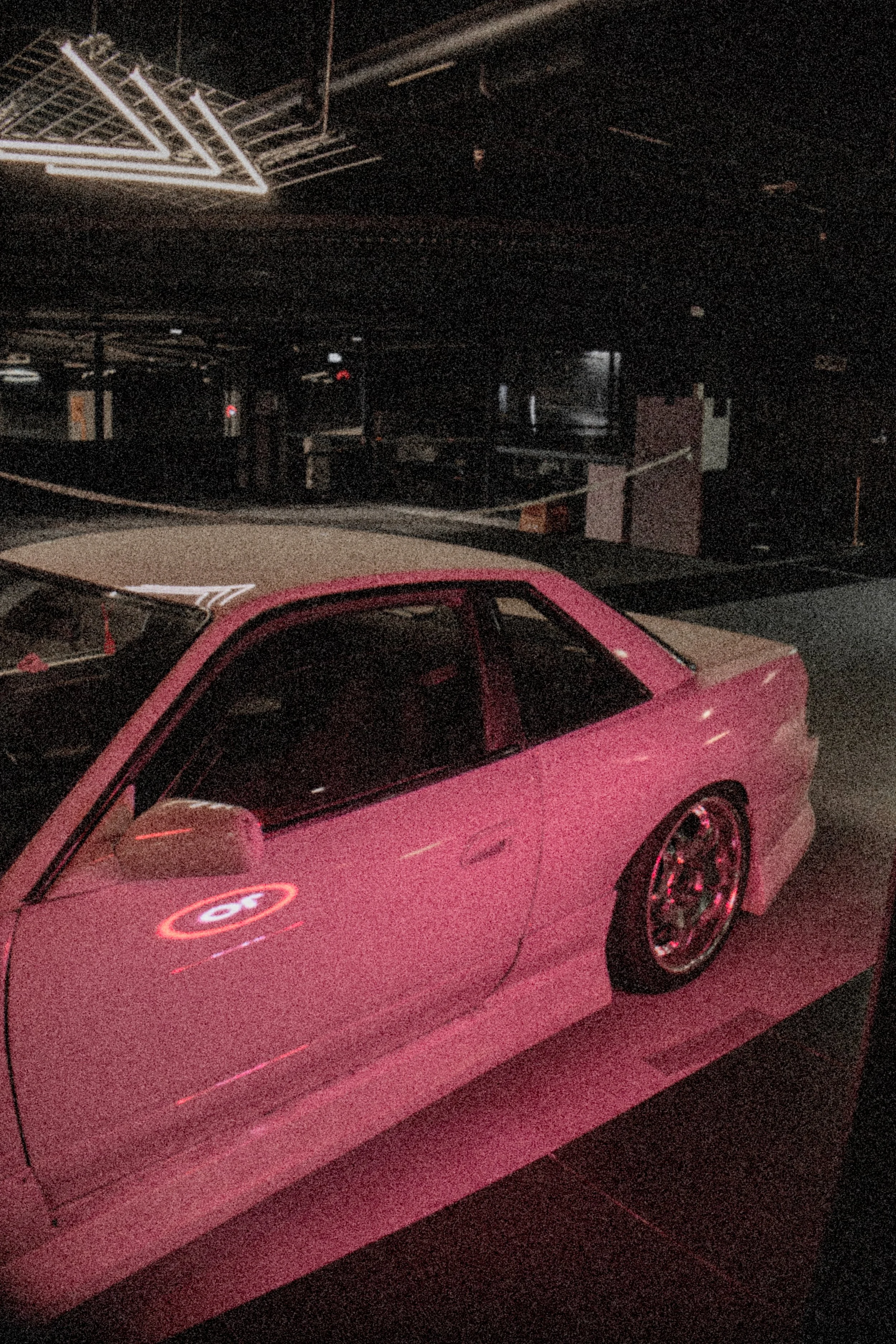 A pink car with stock rims parked inside a dark, dimly lit room with neon lighting reflections.
