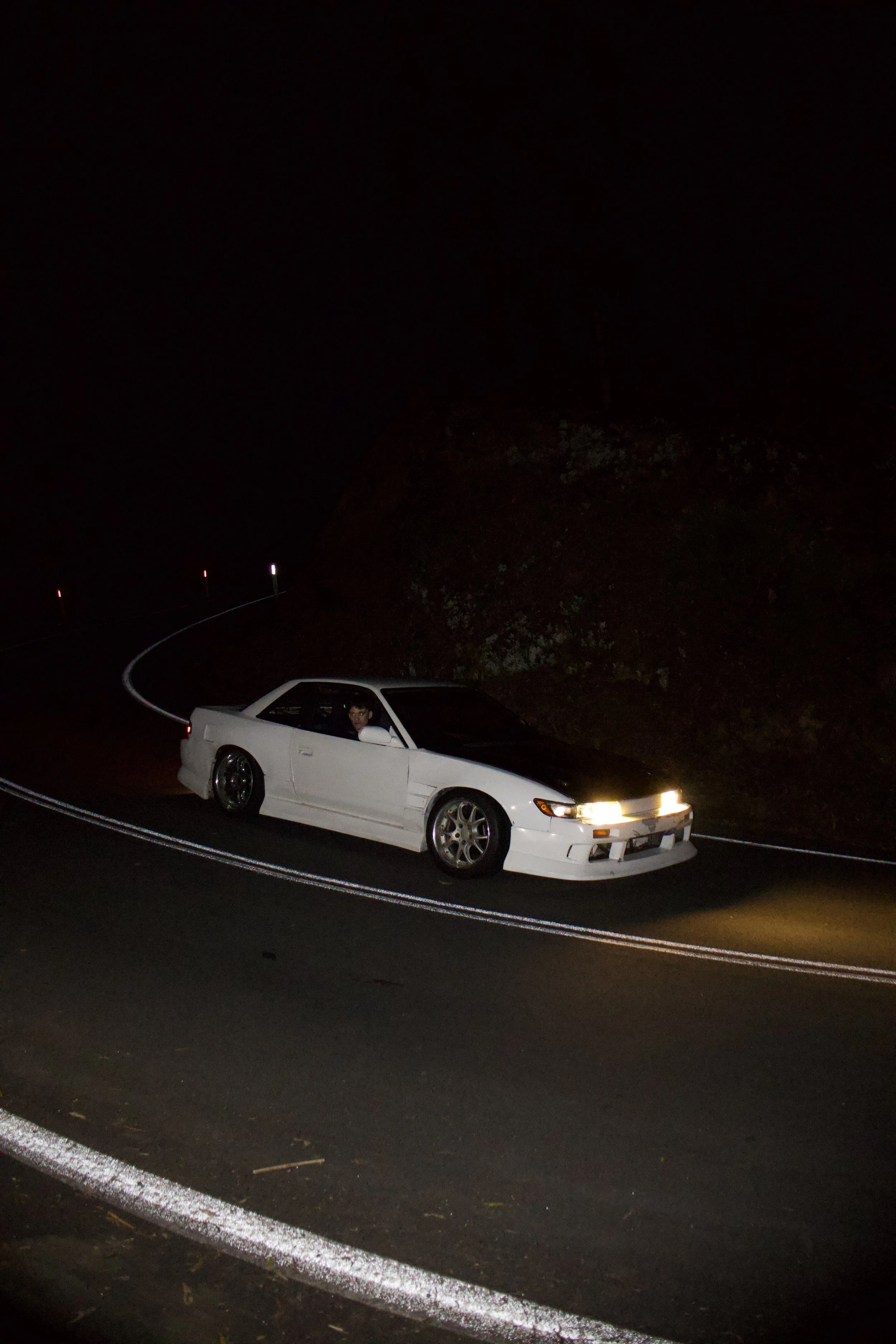 A white car with black hood parked on a dark winding road at night, with a person visible in the driver’s seat.