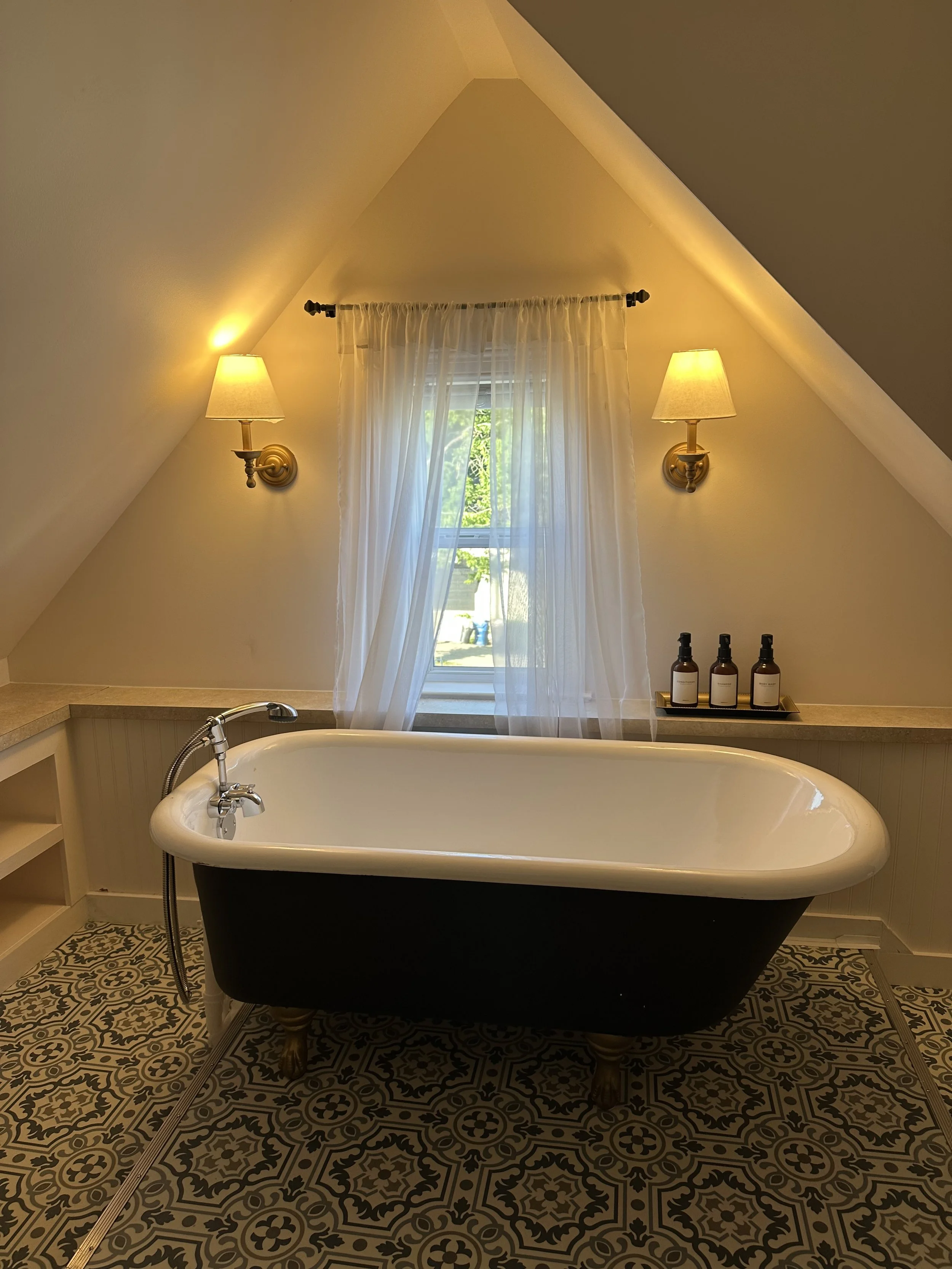 A bathtub in a bathroom with a window, sheer curtains, two wall-mounted lamps, and patterned tile flooring.