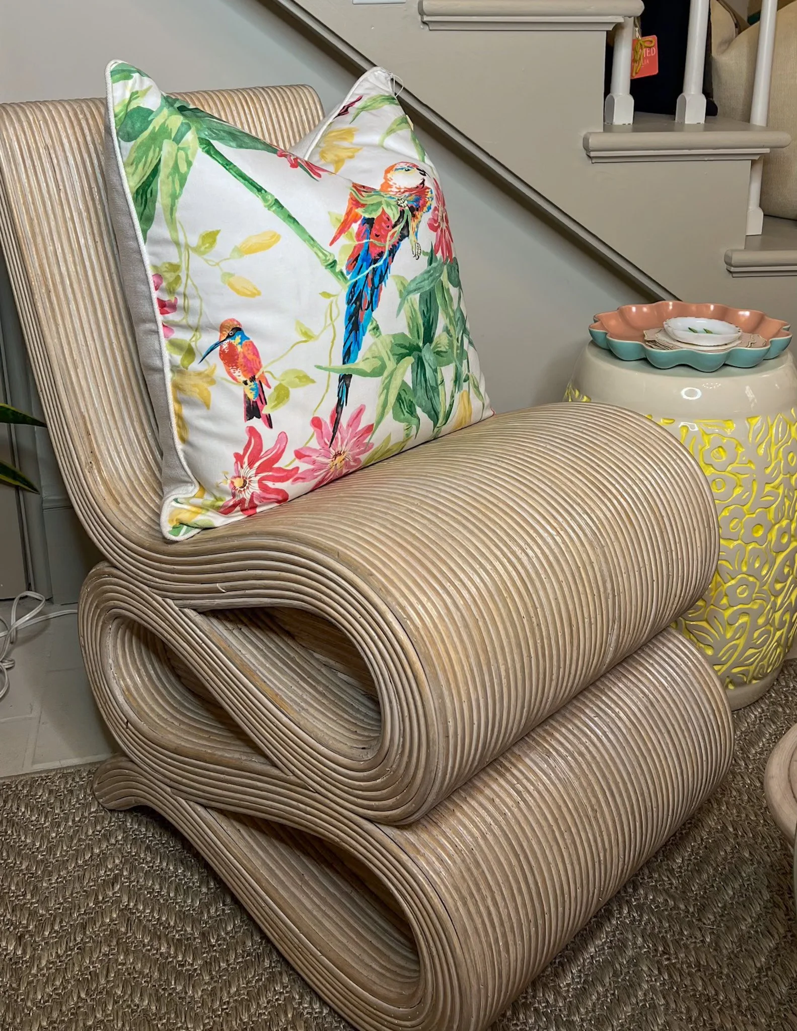 A beige, ribbed, foldable lounge chair with a decorative pillow featuring colorful parrots and foliage. Next to the chair is a yellow and white patterned side table with a pink and teal scalloped dish on top.
