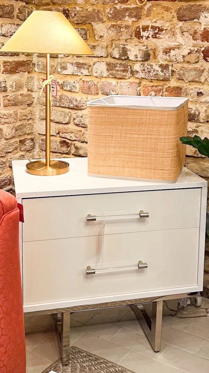 White modern dresser with three drawers, chrome handles, and a decorative clear acrylic bar on the middle drawer, placed against a brick wall. On top of the dresser is a wooden storage box with a white lid and a gold table lamp with a beige shade. A 